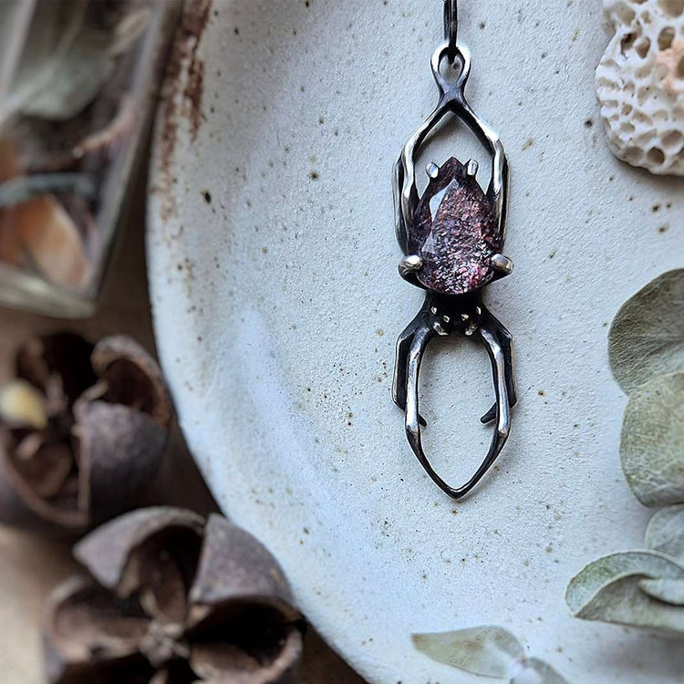 Handcrafted oxidized silver spider pendant with a sparkling purple gemstone on a neutral ceramic background. Nature-inspired gothic jewelry design.