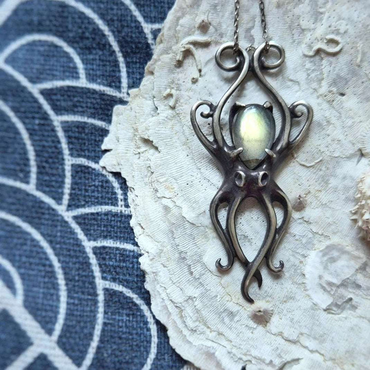 Handmade silver Octopus necklace with glowing labradorite gemstone, photographed on textured shell and indigo fabric background with ethnic pattern