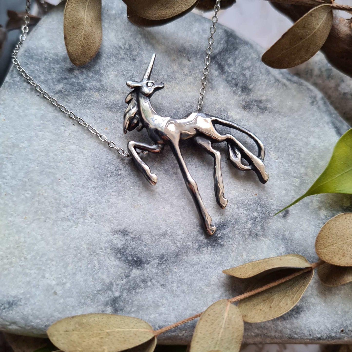 Silver Unicorn necklace placed on smooth stone with dried leaves, evoking a magical forest atmosphere.