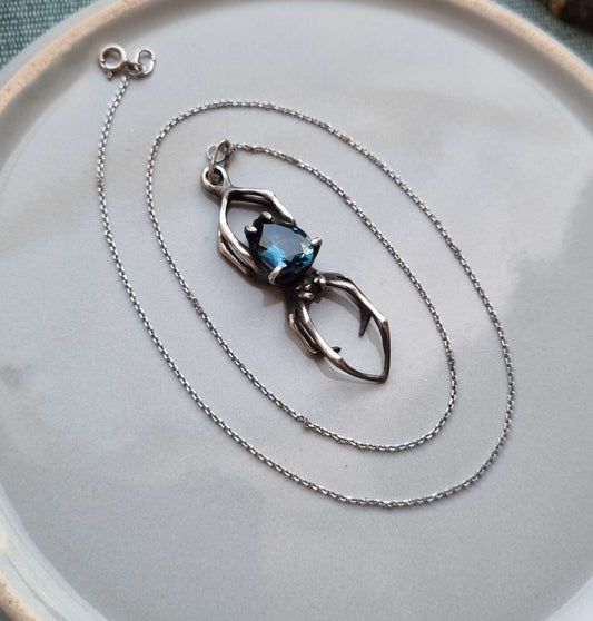 Silver Spider necklace with London blue topaz laid on a ceramic plate, styled with a silver chain in an oval spiral