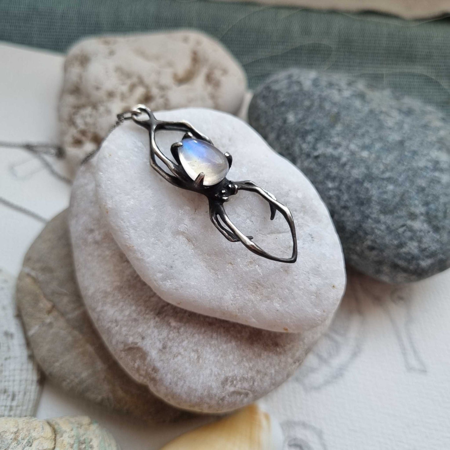 Silver spider necklace with rainbow moonstone placed on smooth stones, styled with beach pebbles