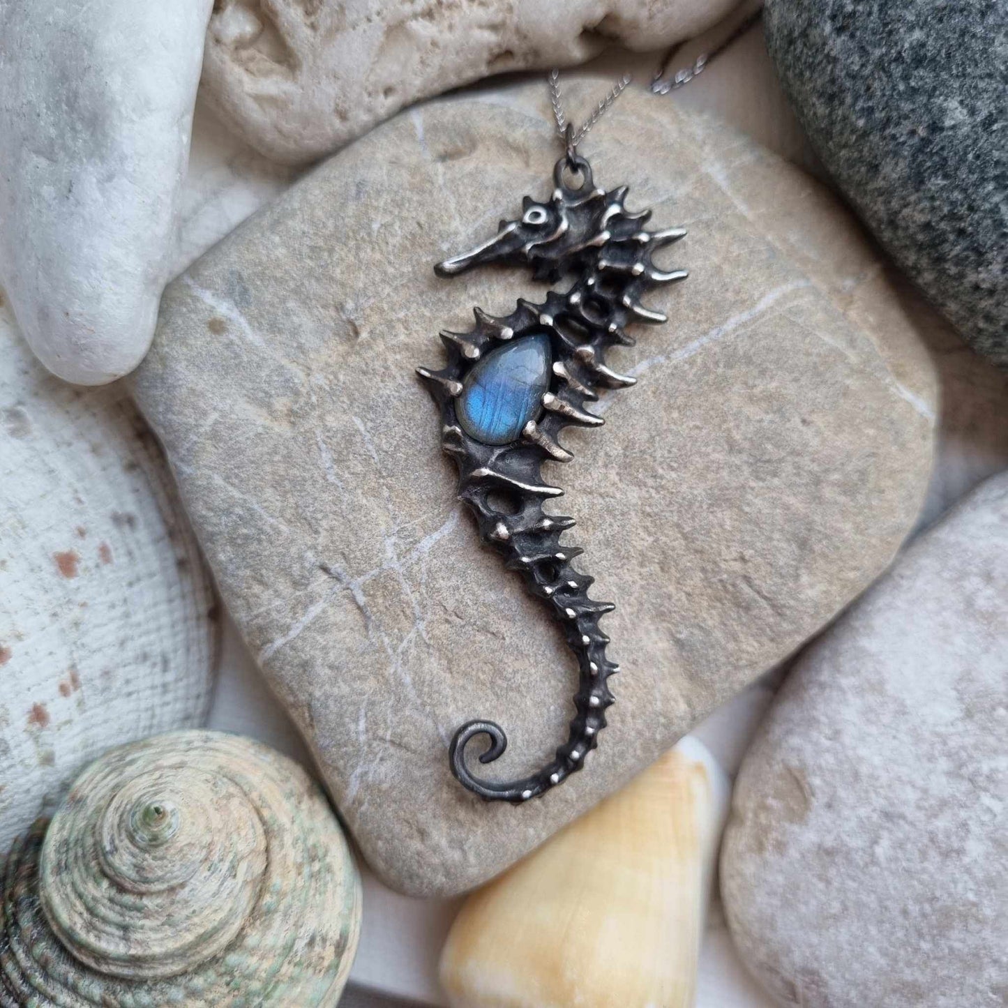 Handmade silver Seahorse necklace with labradorite gemstone resting on a stone with seashells around.