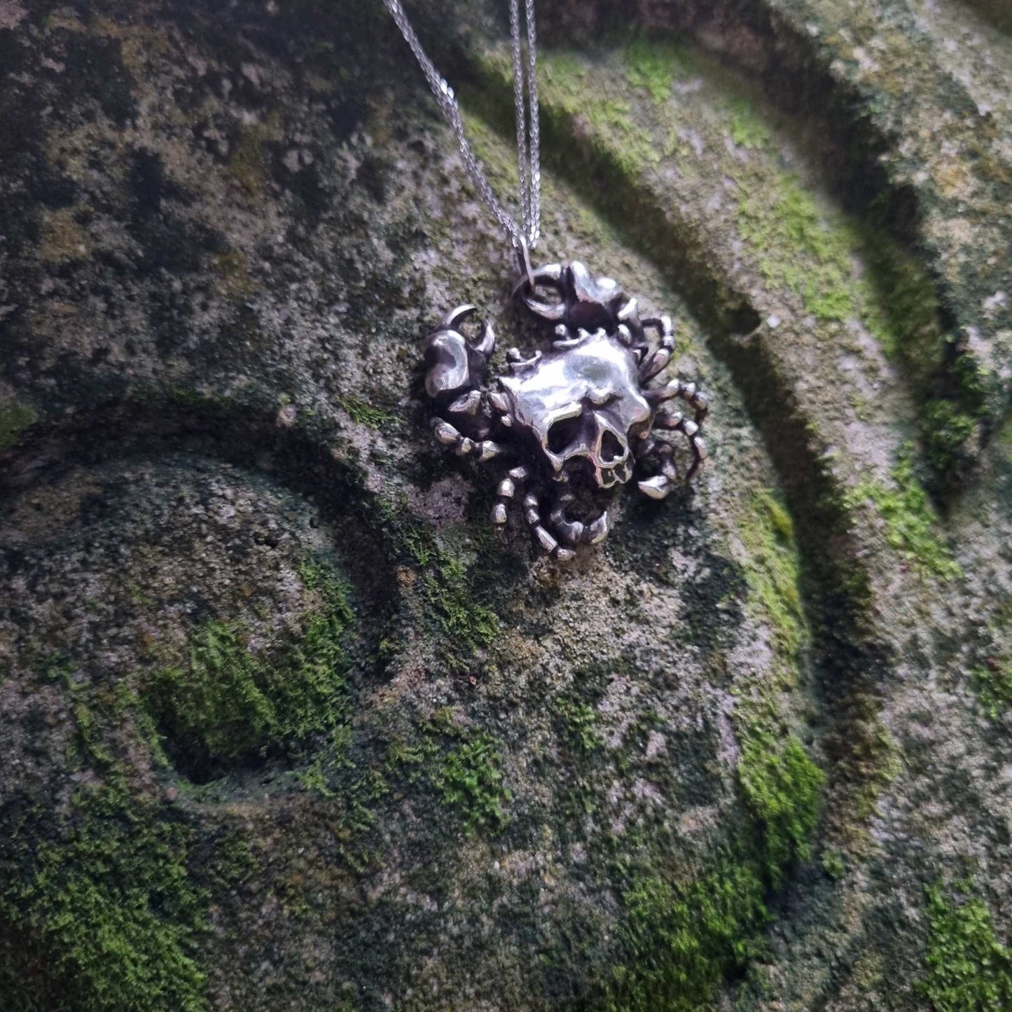 Silver Skull necklace with detailed crab legs and a skull, lying on a textured green mossy rock with a spiral pattern