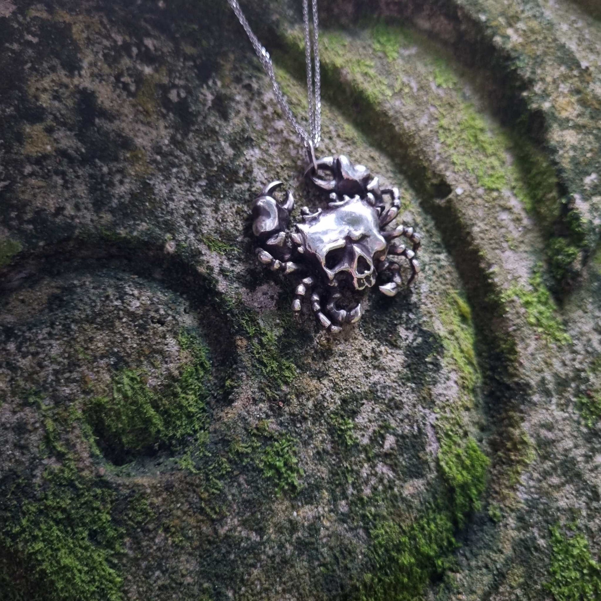 Silver Skull necklace with detailed crab legs and a skull, lying on a textured green mossy rock with a spiral pattern