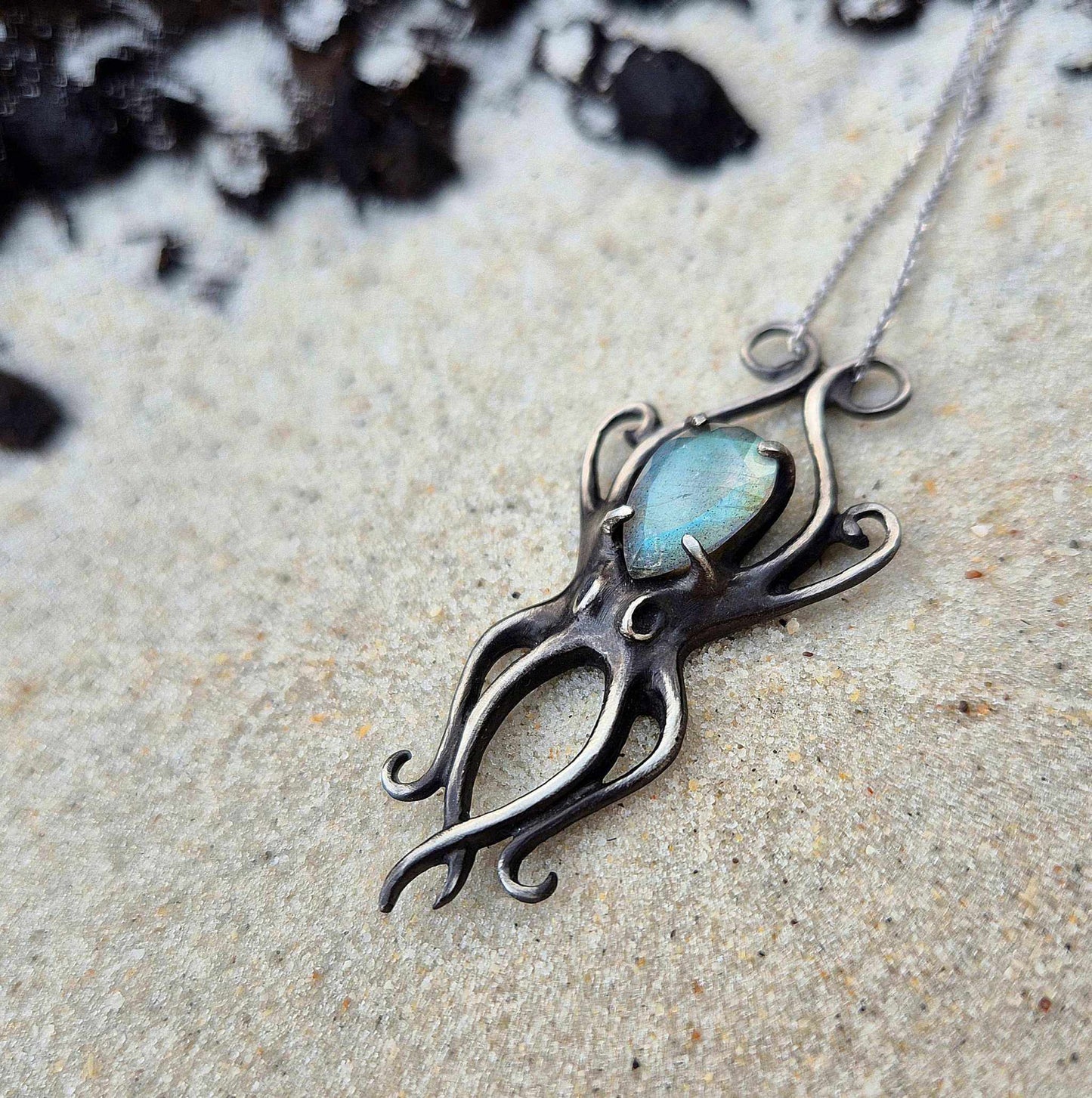 Silver Octopus necklace with blue labradorite displayed on sand with dark rocks nearby