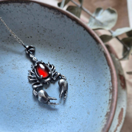 Silver Scorpion necklace with a vivid red garnet gemstone displayed on a pale blue plate with dry botanical accents in the background.