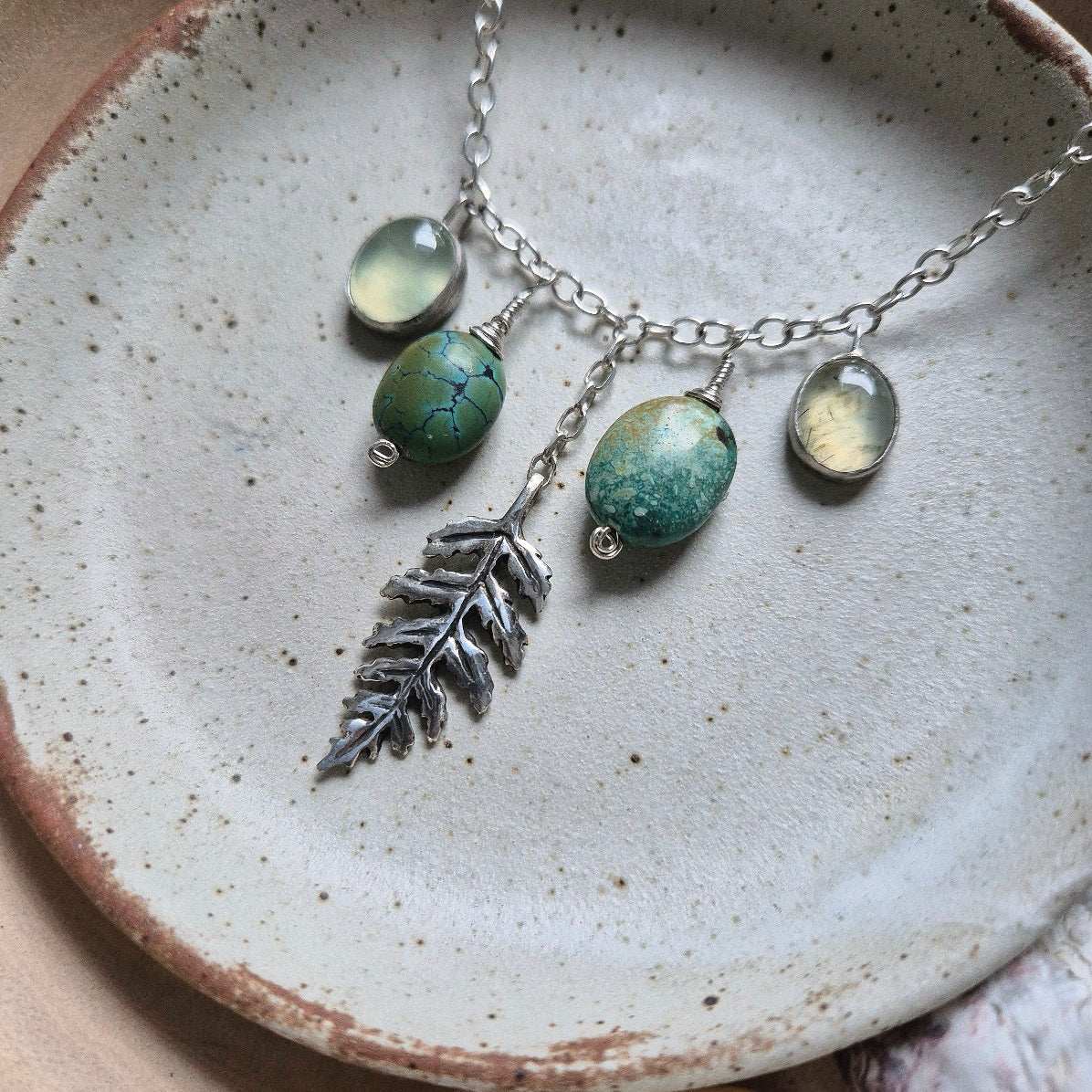 Close-up of forest-inspired silver turquoise necklace with turquoise beads, prehnite cabochons, and a sculpted leaf charm, displayed on a handmade ceramic dish