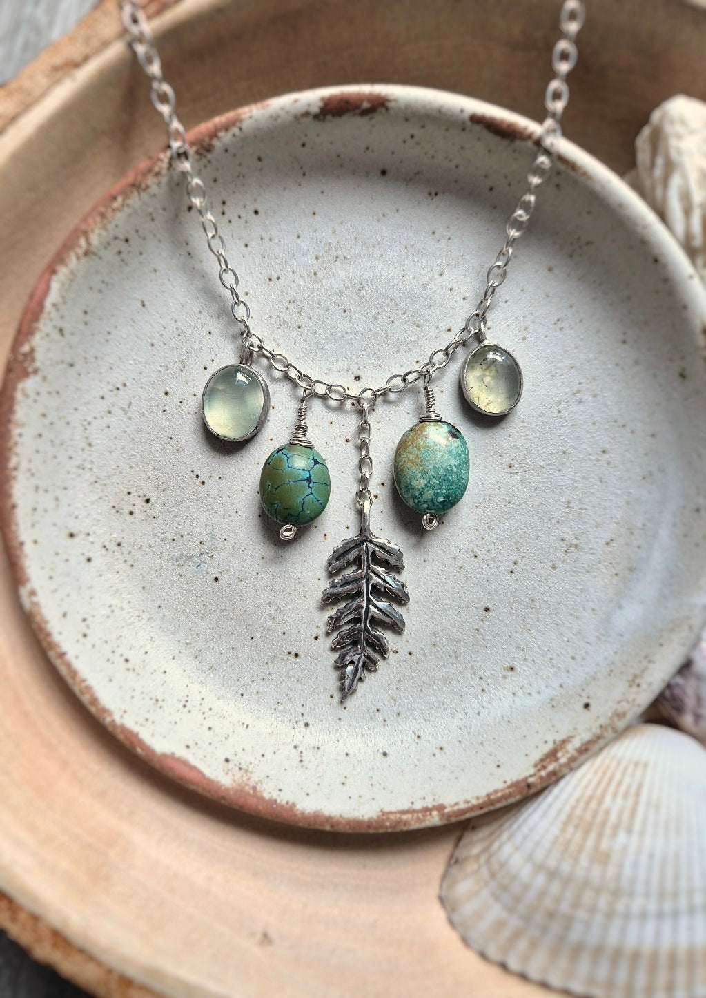 Flat lay of Forest path turquoise necklace featuring turquoise, prehnite, and a sculptural silver leaf, styled with shells and ceramic for a natural coastal touch.
