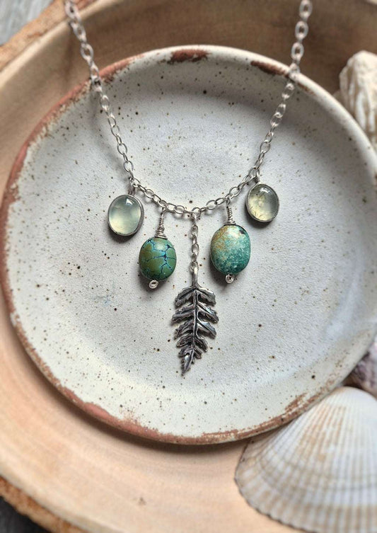 Flat lay of Forest path turquoise necklace featuring turquoise, prehnite, and a sculptural silver leaf, styled with shells and ceramic for a natural coastal touch.