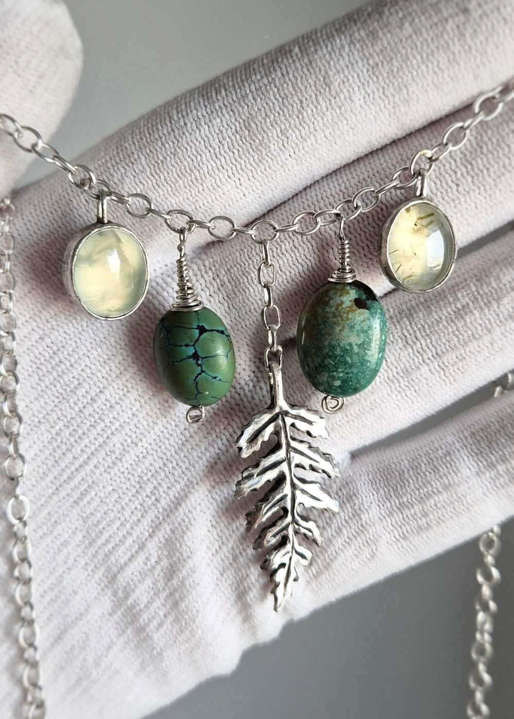 Close-up of handcrafted silver turquoise  necklace with turquoise and prehnite gemstones, highlighting textures and details against a soft fabric background.