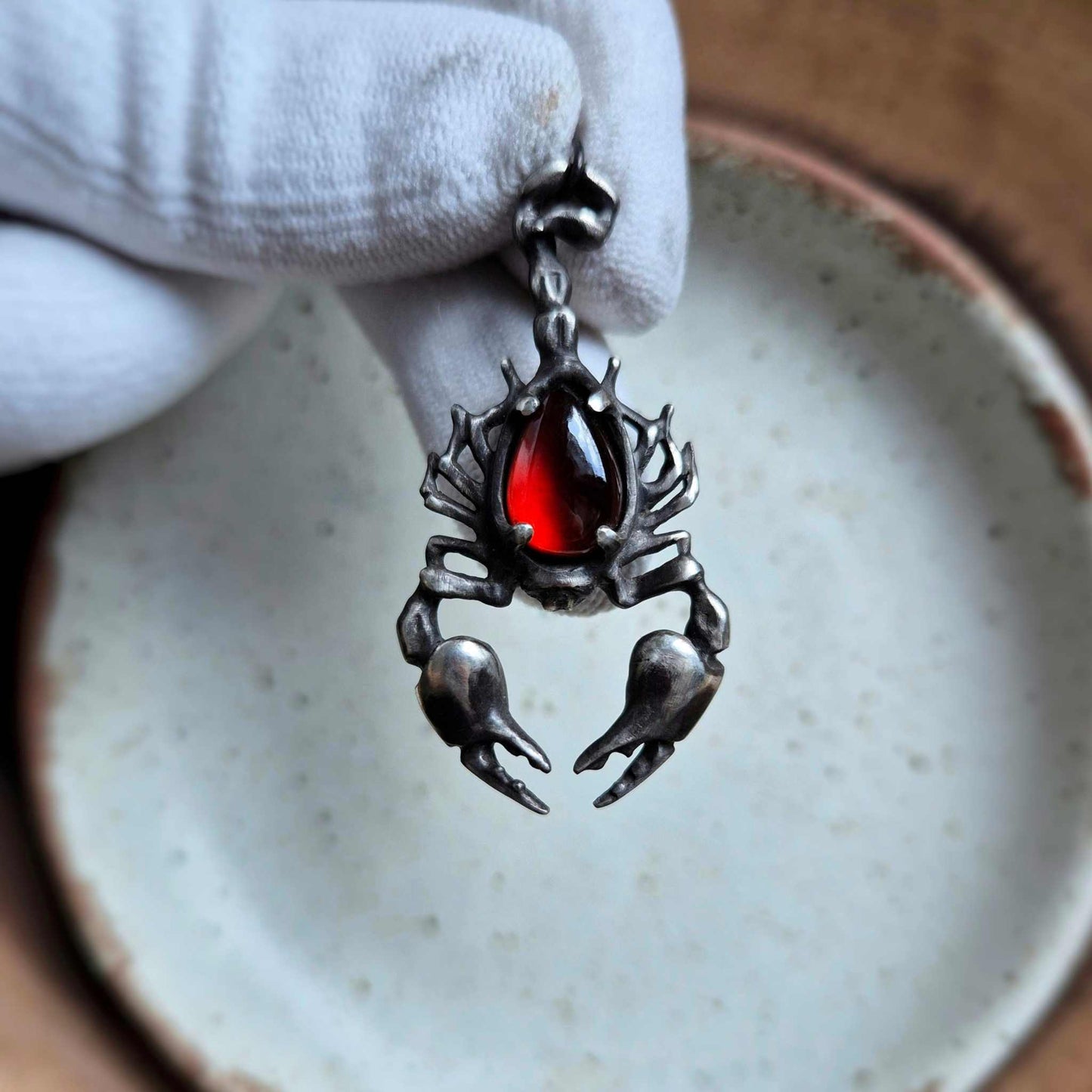 Gloved hand holding a silver Scorpion necklace with a deep red stone, placed above a softly speckled plate