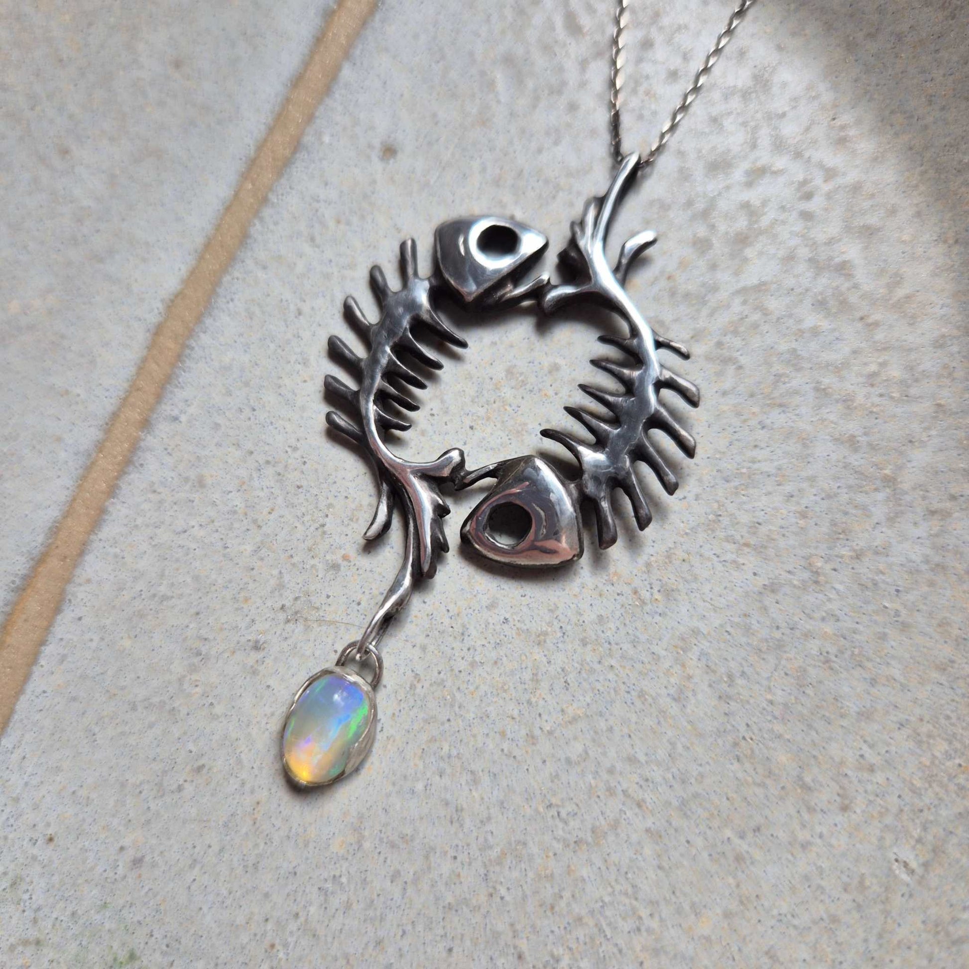Front view of silver fish skeleton necklace with glowing opal drop. Pisces-inspired talisman pendant on minimalist background