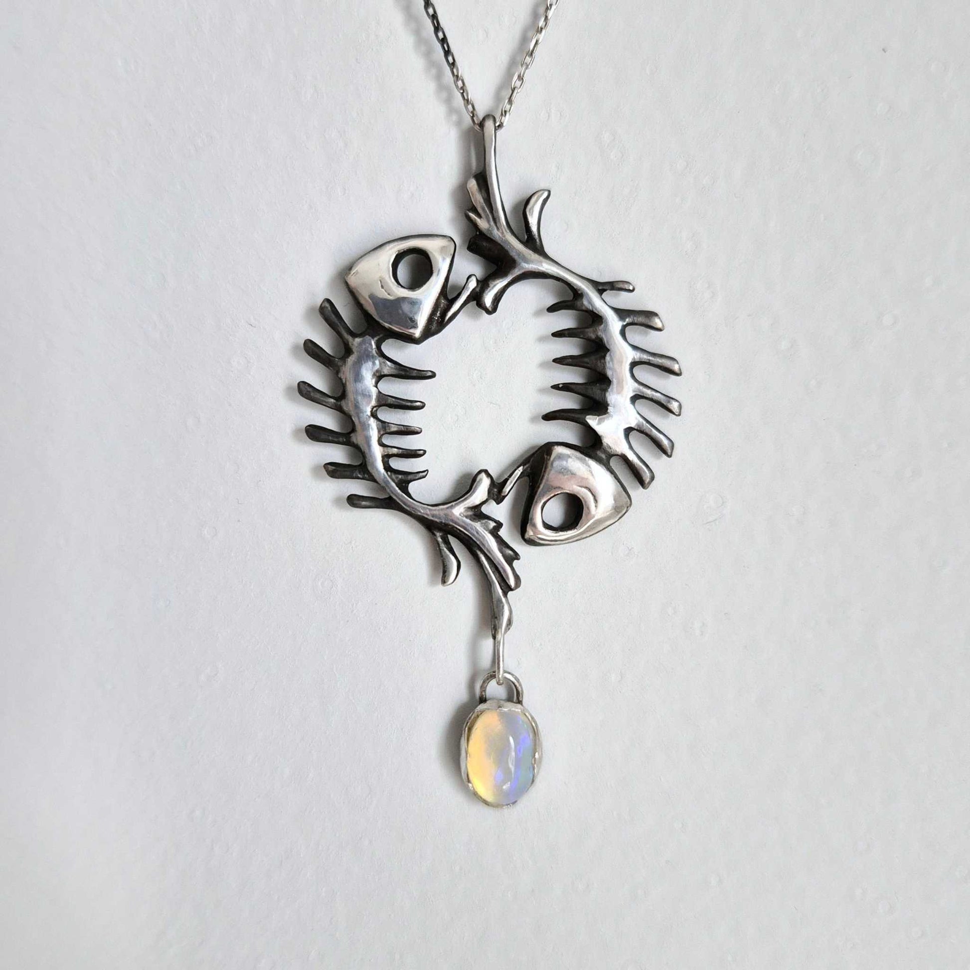 Handmade silver fish skeleton necklace with oval opal, shown on ceramic surface. Symbolic pendant featuring circular fishbone design.
