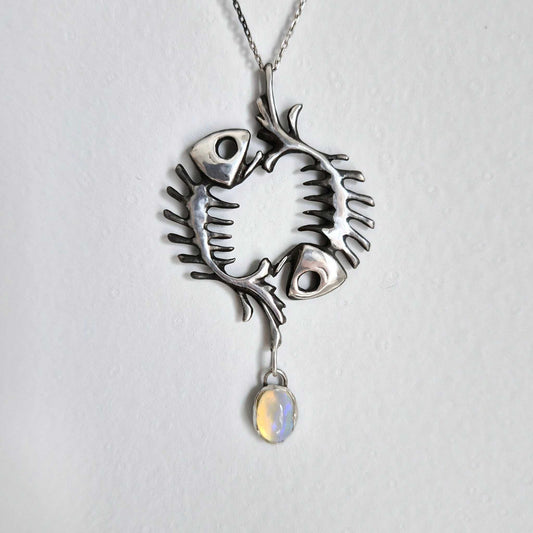 Handmade silver fish skeleton necklace with oval opal, shown on ceramic surface. Symbolic pendant featuring circular fishbone design.