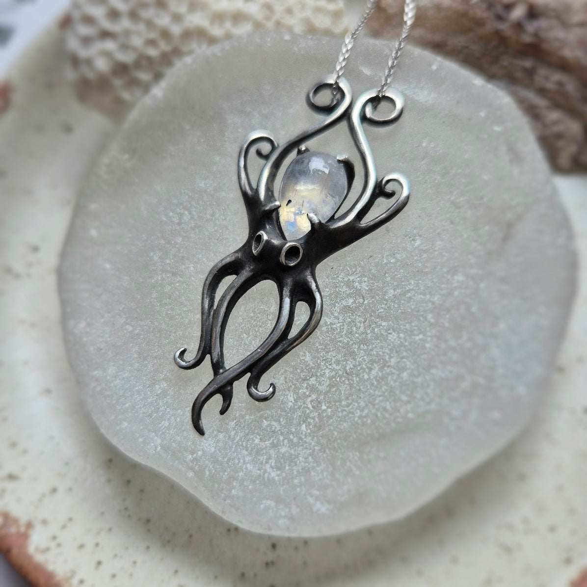 Textured sterling silver Octopus necklace with shimmering rainbow moonstone, laid on rustic ceramic surface.