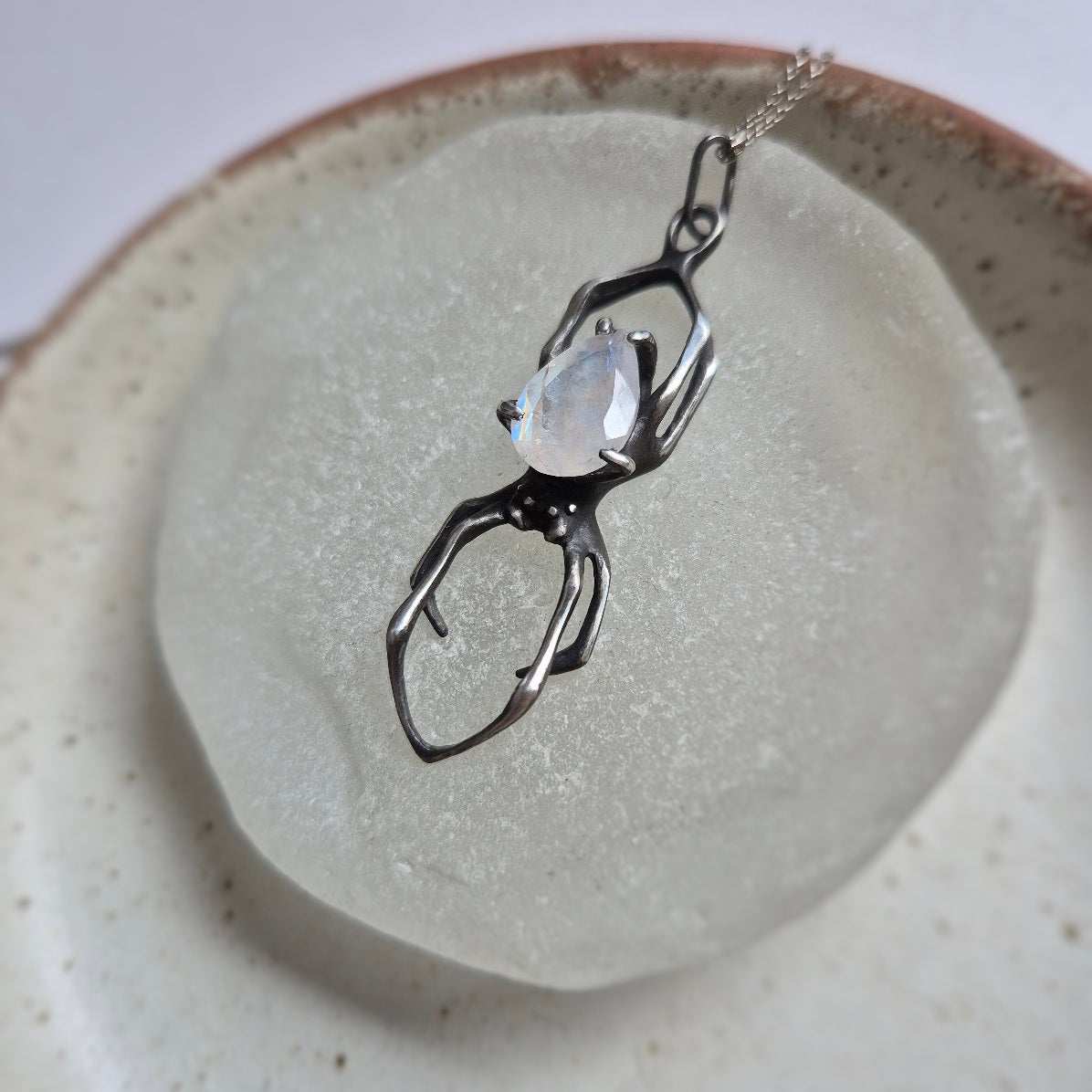 Silver Spider necklace with rainbow moonstone resting on ceramic surface