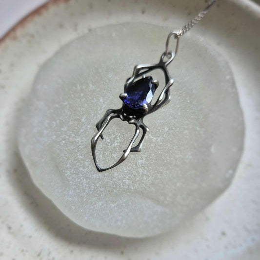 Silver Spider necklace with iolite sunstone displayed on a matte glass plate, showing its intricate legs and dark shimmer