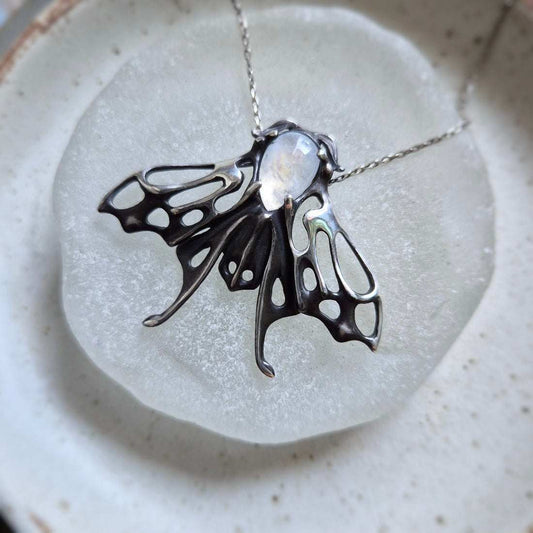 Close-up of a Silver moth necklace with rainbow moonstone displayed on round frosted glass plate.