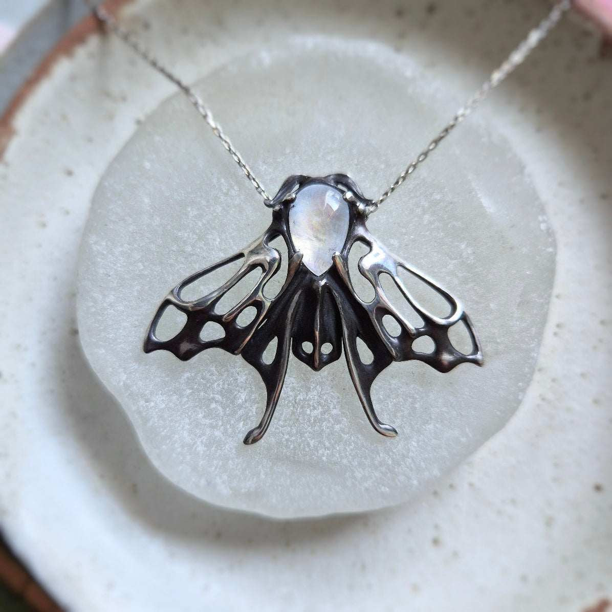 Articulated silver moth pendant with moonstone, resting on circular white backdrop