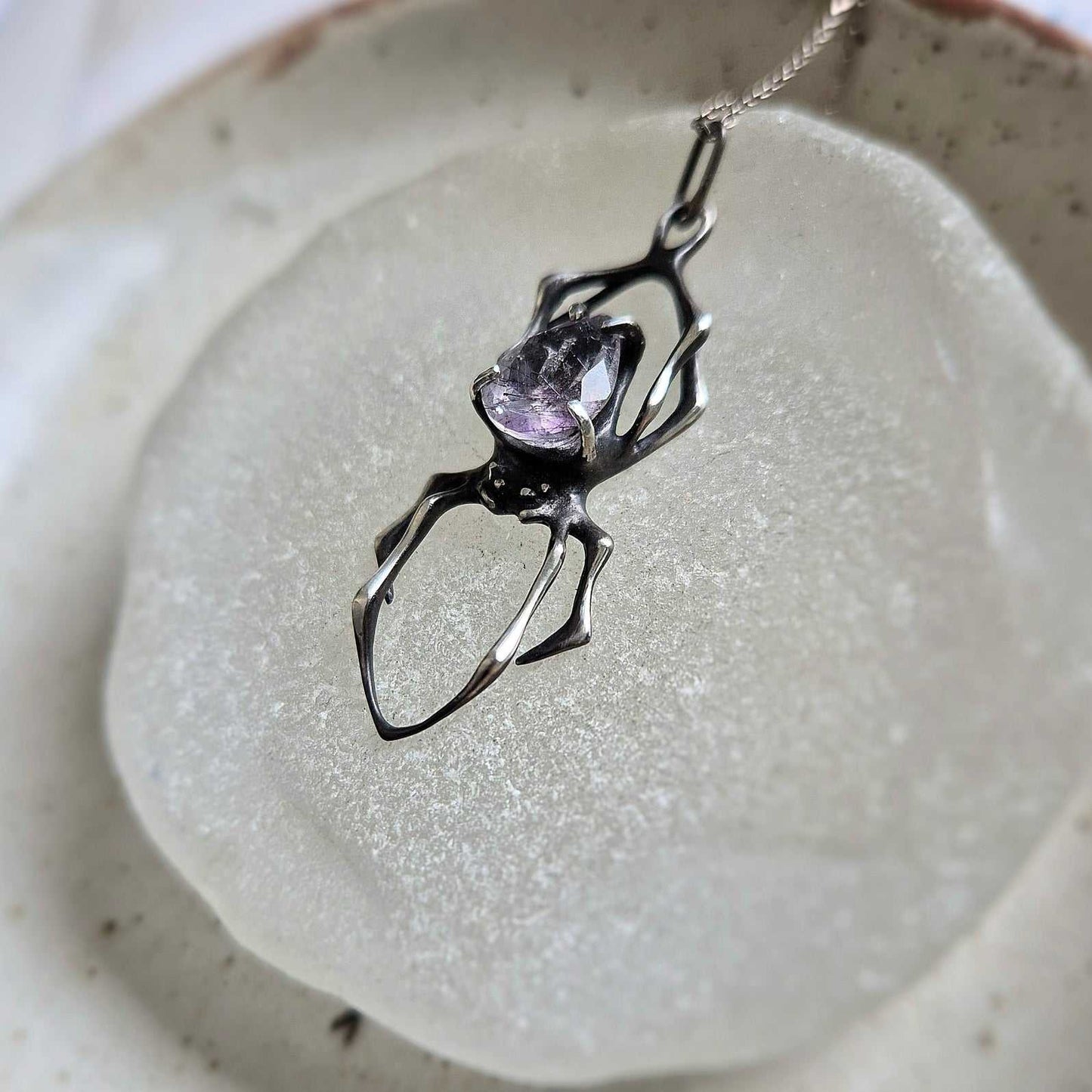 Silver Spider necklace with Super Seven gemstone resting on a frosted glass surface, showing the pendant’s sculptural openwork design.