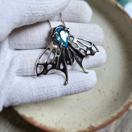 Hand holding the silver Moth pendant with vivid blue topaz — details of wings and gemstone clearly visible.