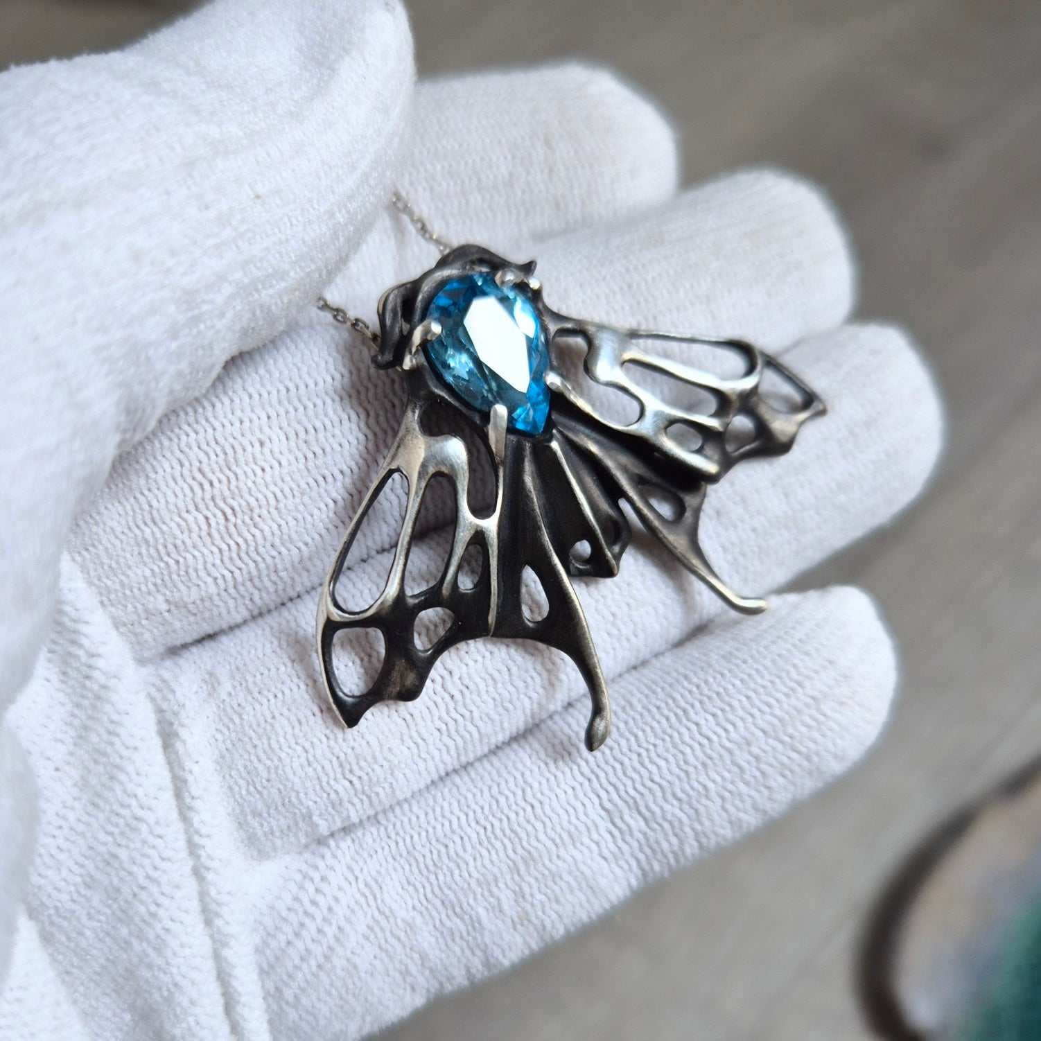 Close-up of the silver Moth necklace in a gloved hand, highlighting the contrast between oxidized silver and the bright blue stone.