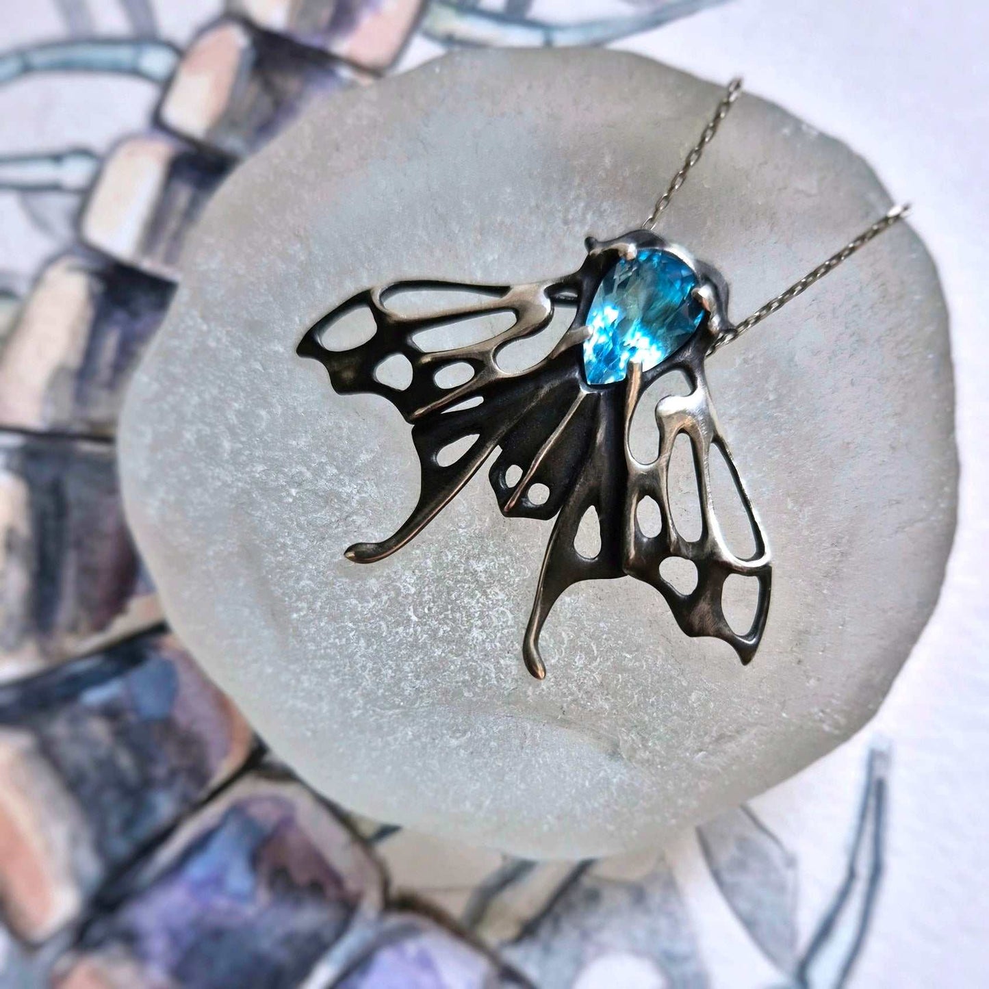 Silver Moth necklace with Swiss blue topaz resting on pale glass with a background of watercolor sketch