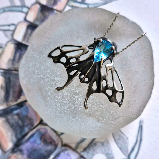 Silver Moth necklace with Swiss blue topaz resting on pale glass with a background of watercolor sketch