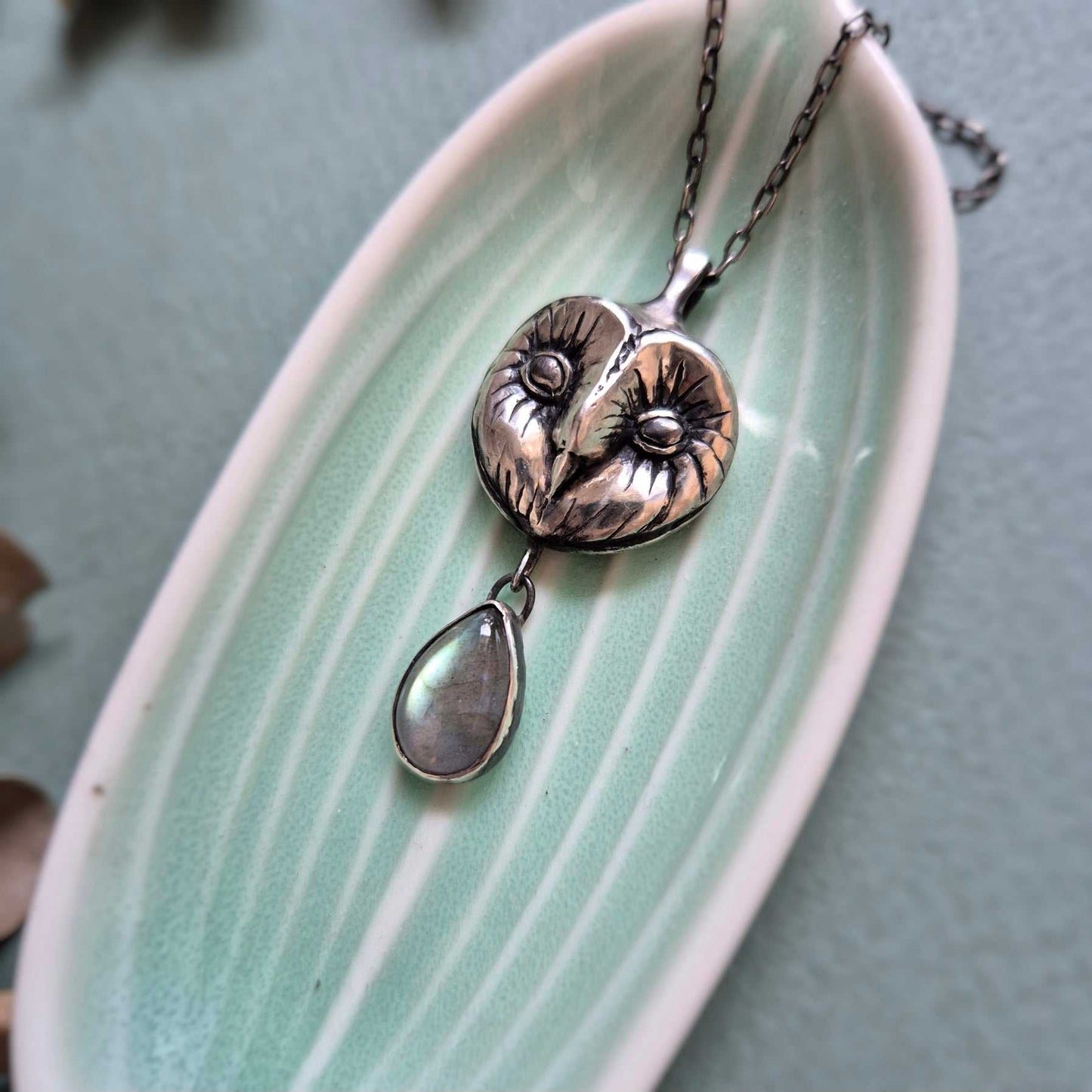 Barn owl–inspired silver Owl necklace lying on a pale green ceramic dish with botanical decor.