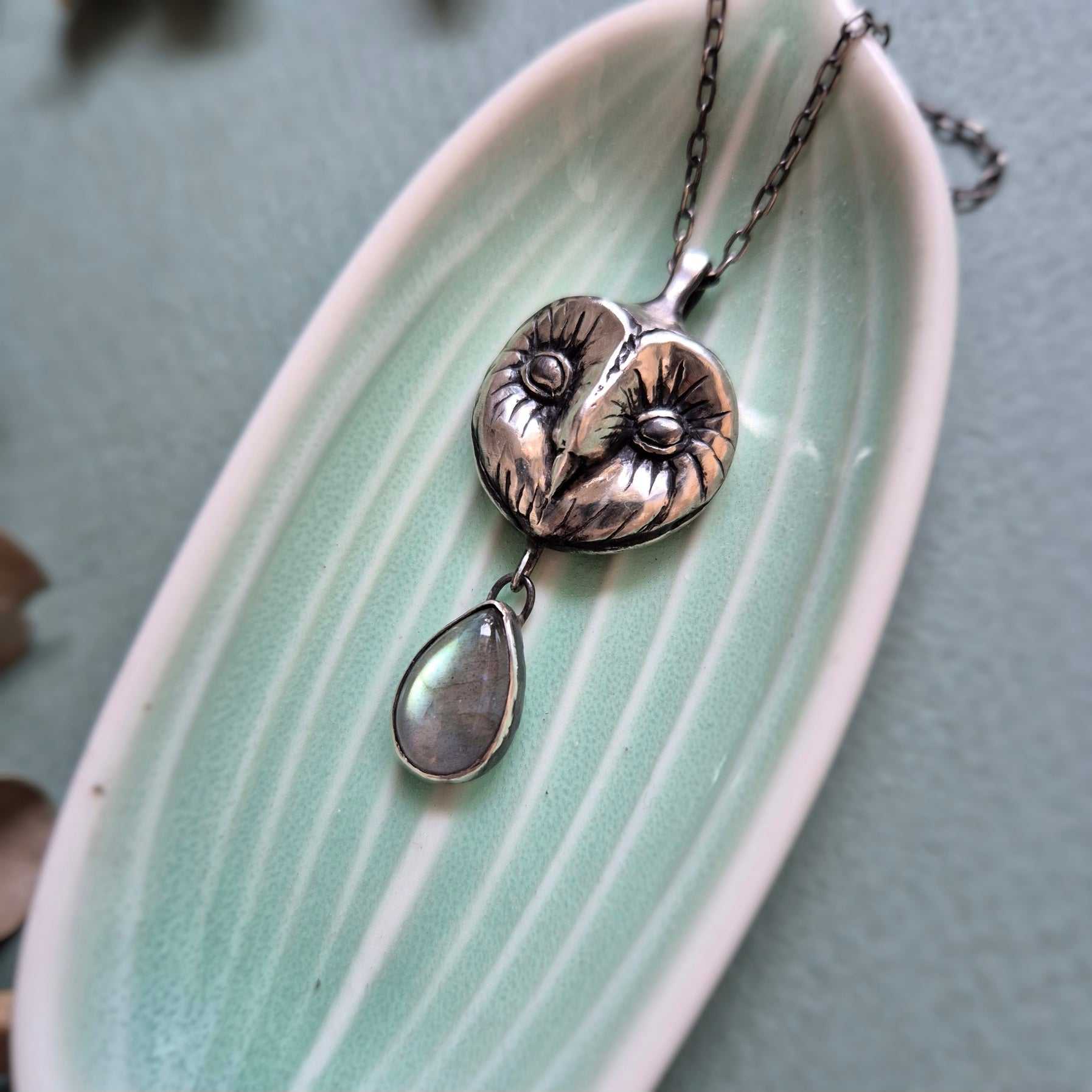 Barn owl–inspired silver Owl necklace lying on a pale green ceramic dish with botanical decor.