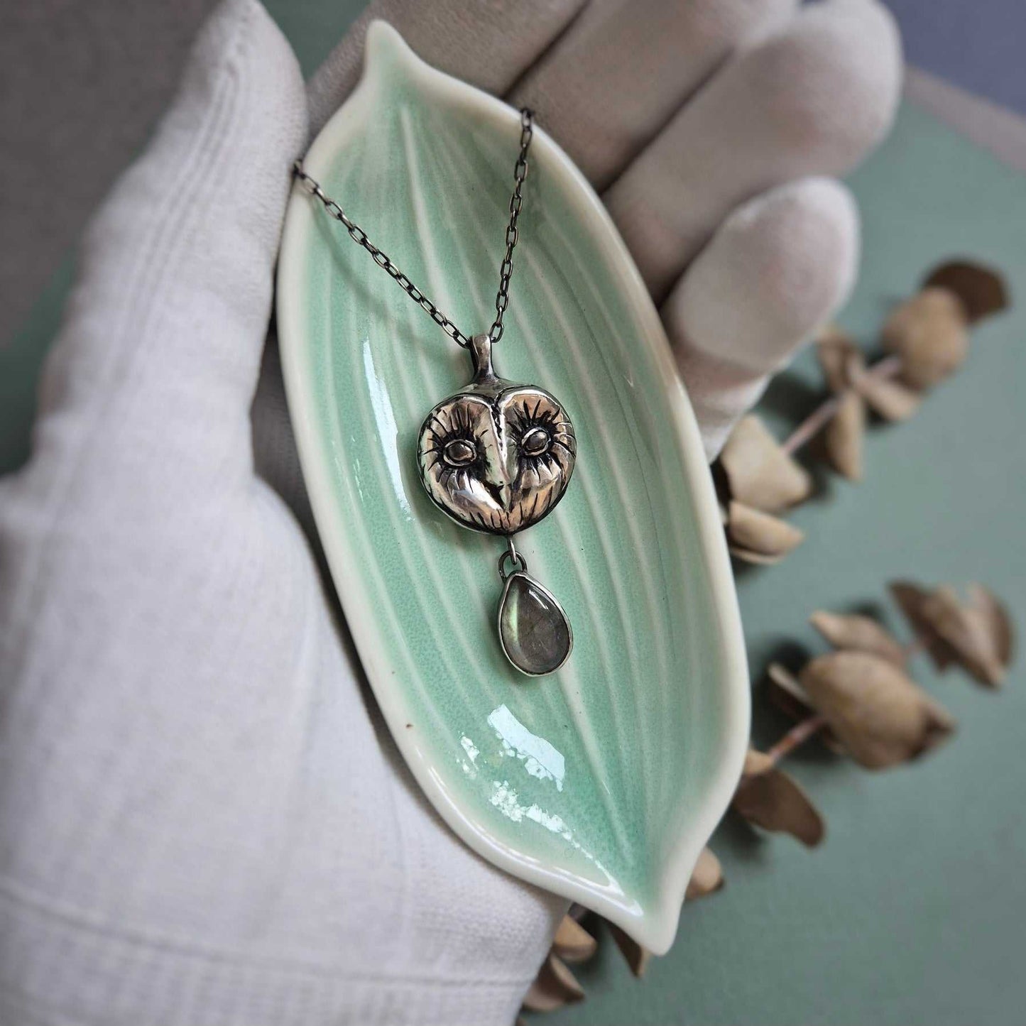 Silver Owl necklace resting on a tiny dish in a gloved hand, showcasing its texture and symbolic charm.