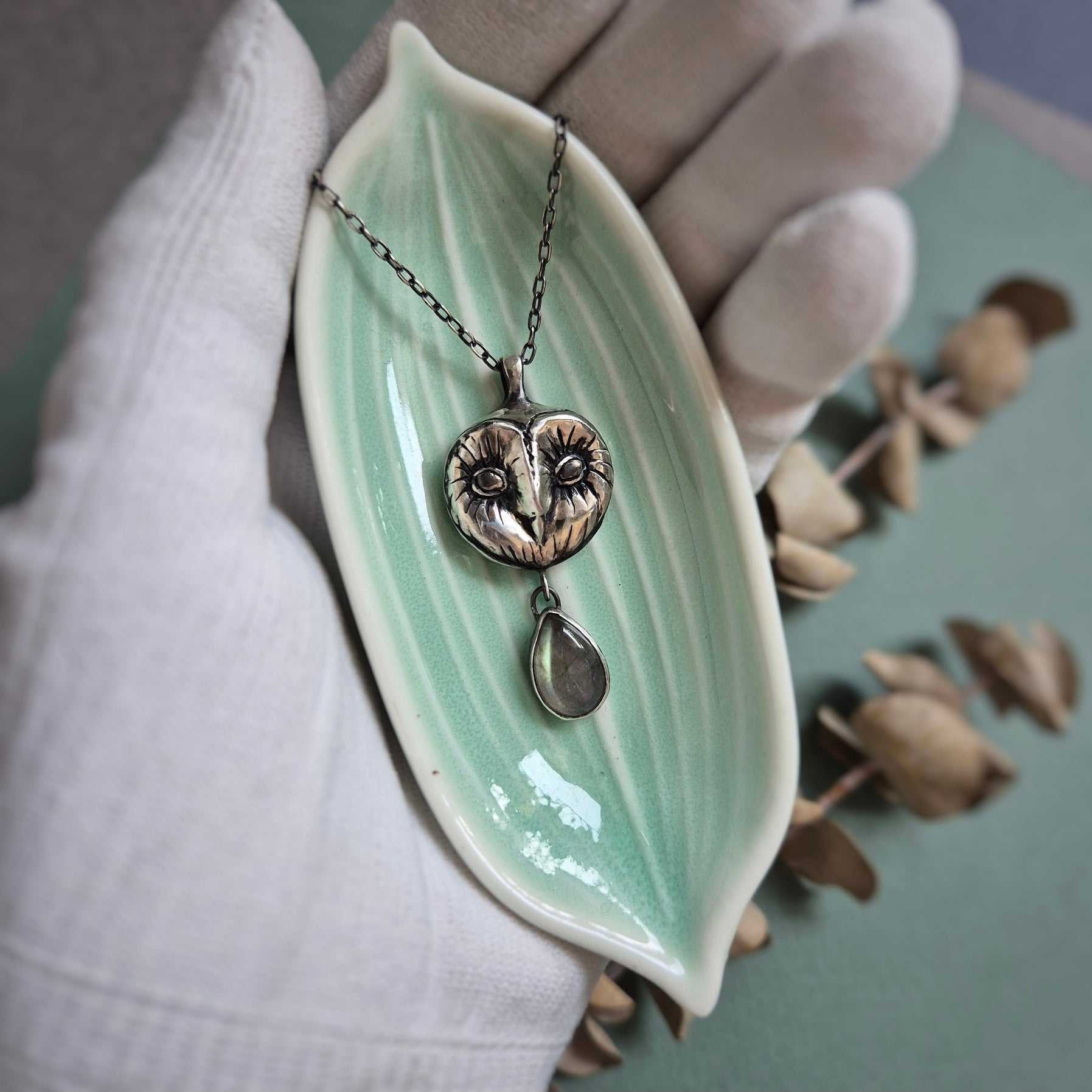 Silver Owl necklace resting on a tiny dish in a gloved hand, showcasing its texture and symbolic charm.