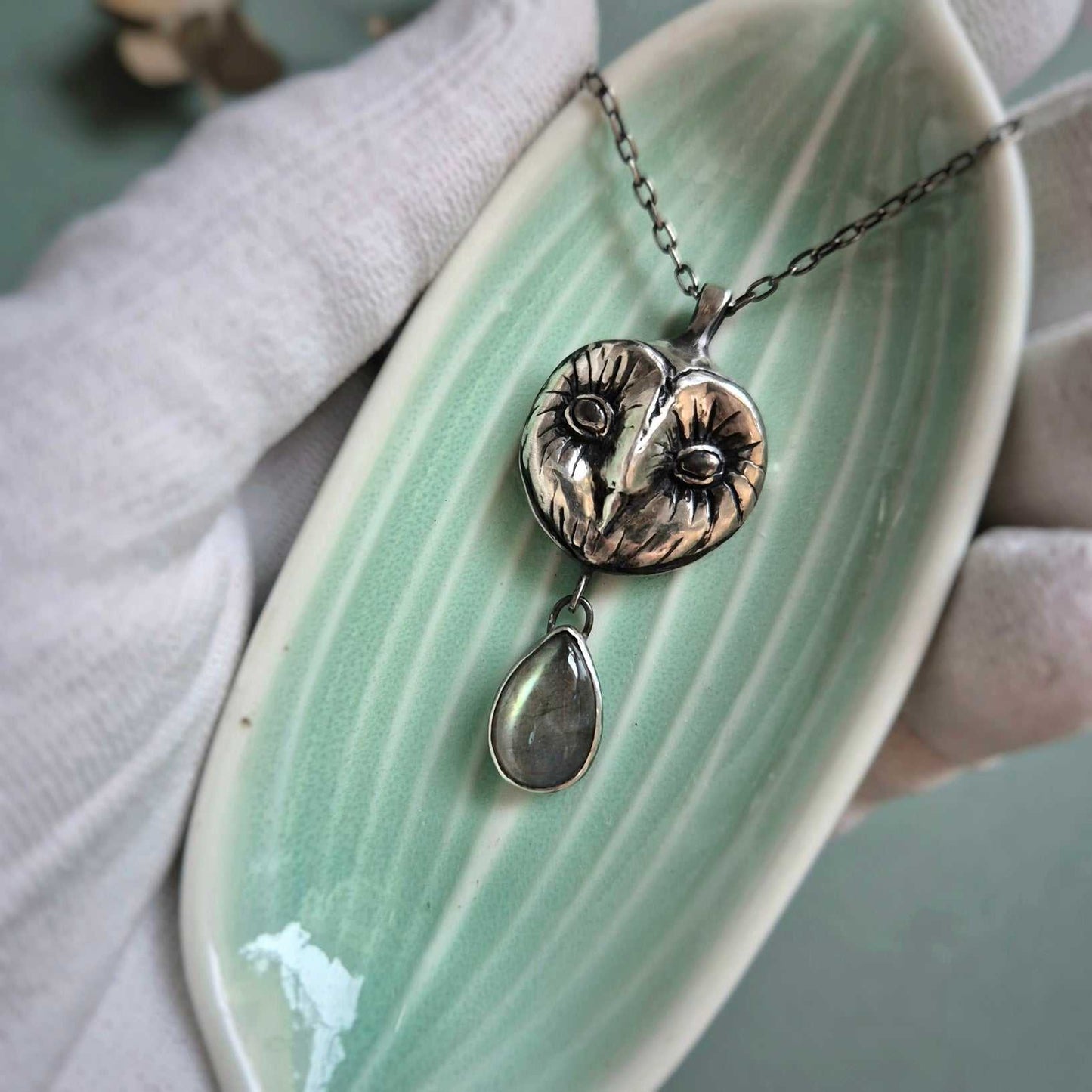 Silver Owl necklace resting on a tiny dish in a gloved hand, showing carved Barn owl face and a gem drop