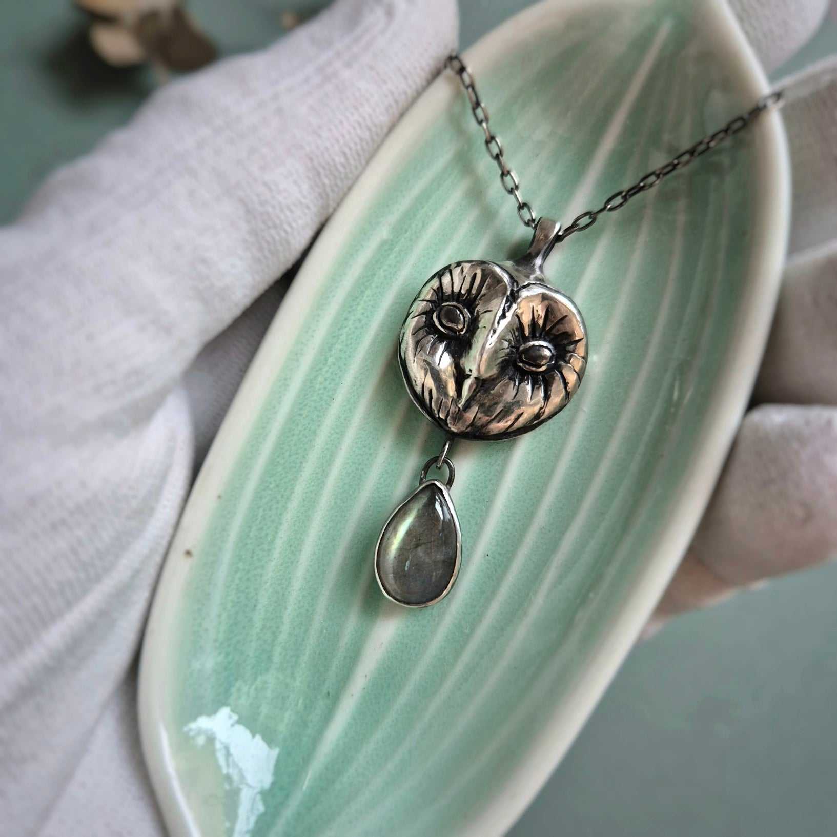 Silver Owl necklace resting on a tiny dish in a gloved hand, showing carved Barn owl face and a gem drop