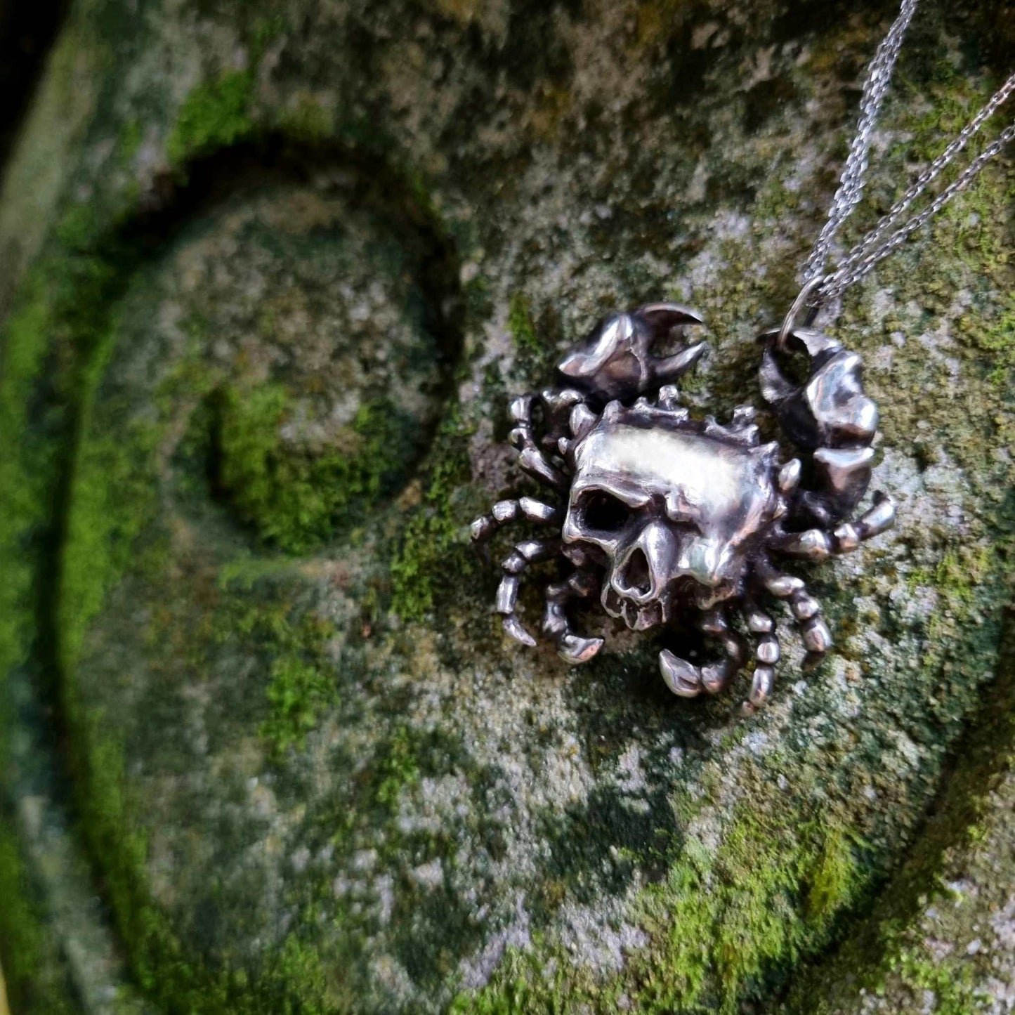 Silver Skull necklace photographed on moss-covered stone, highlighting its crab-like claws and eerie skull detail.