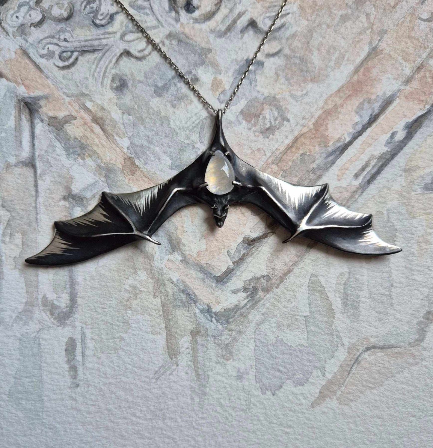 Sculptural silver Bat necklace with wide wings and glowing moonstone, on watercolor sketch