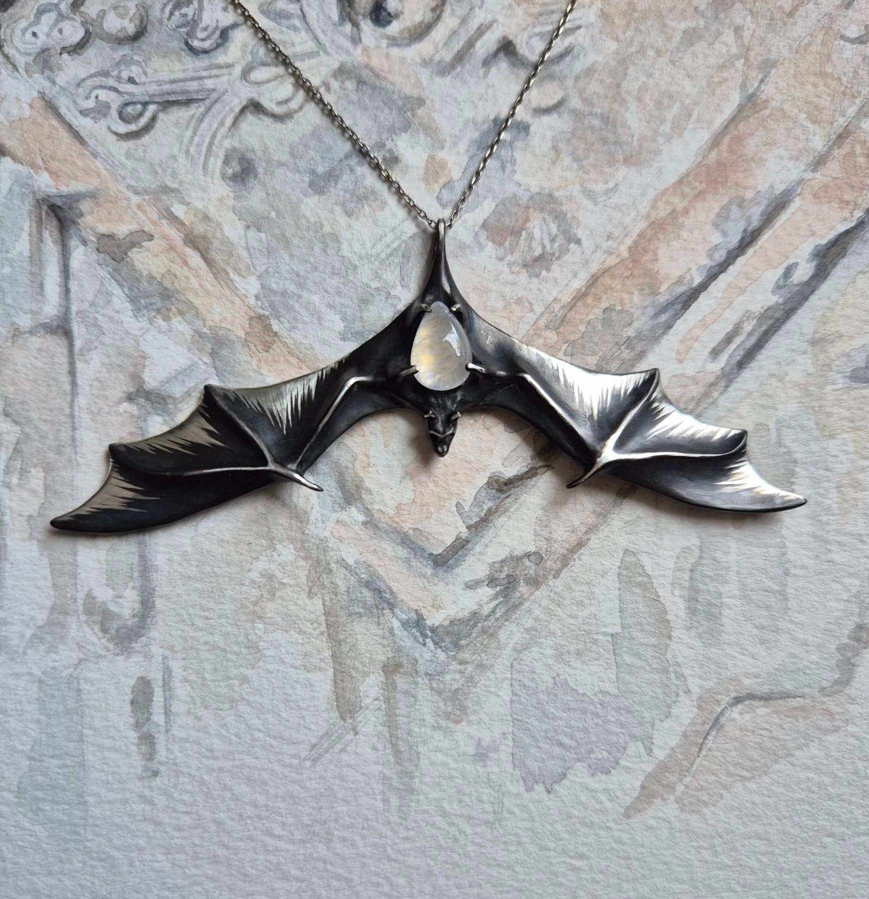 Sculptural silver Bat necklace with wide wings and glowing moonstone, on watercolor sketch