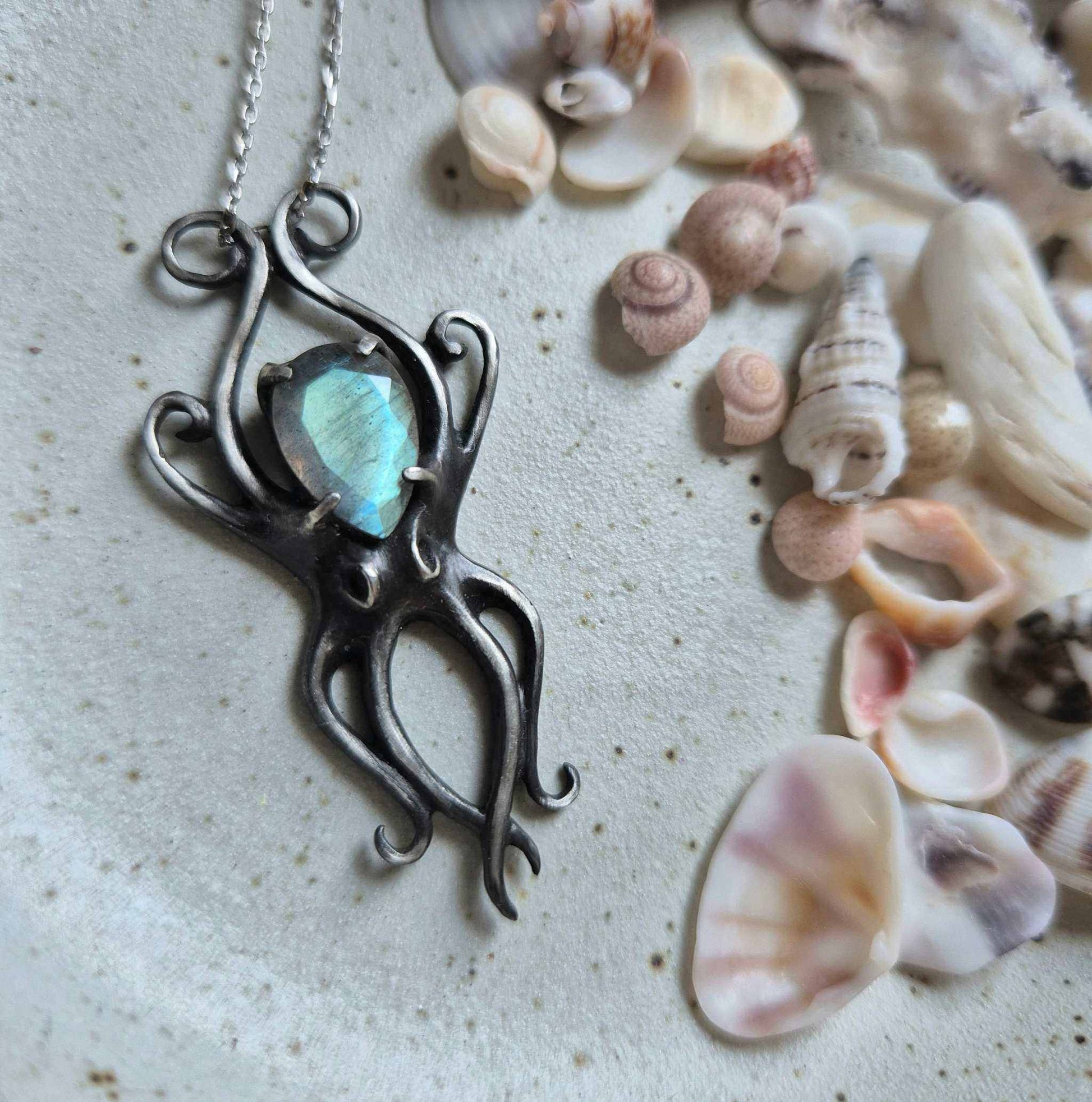 Close-up of silver Octopus pendant with cyan labradorite on ceramic background with seashells