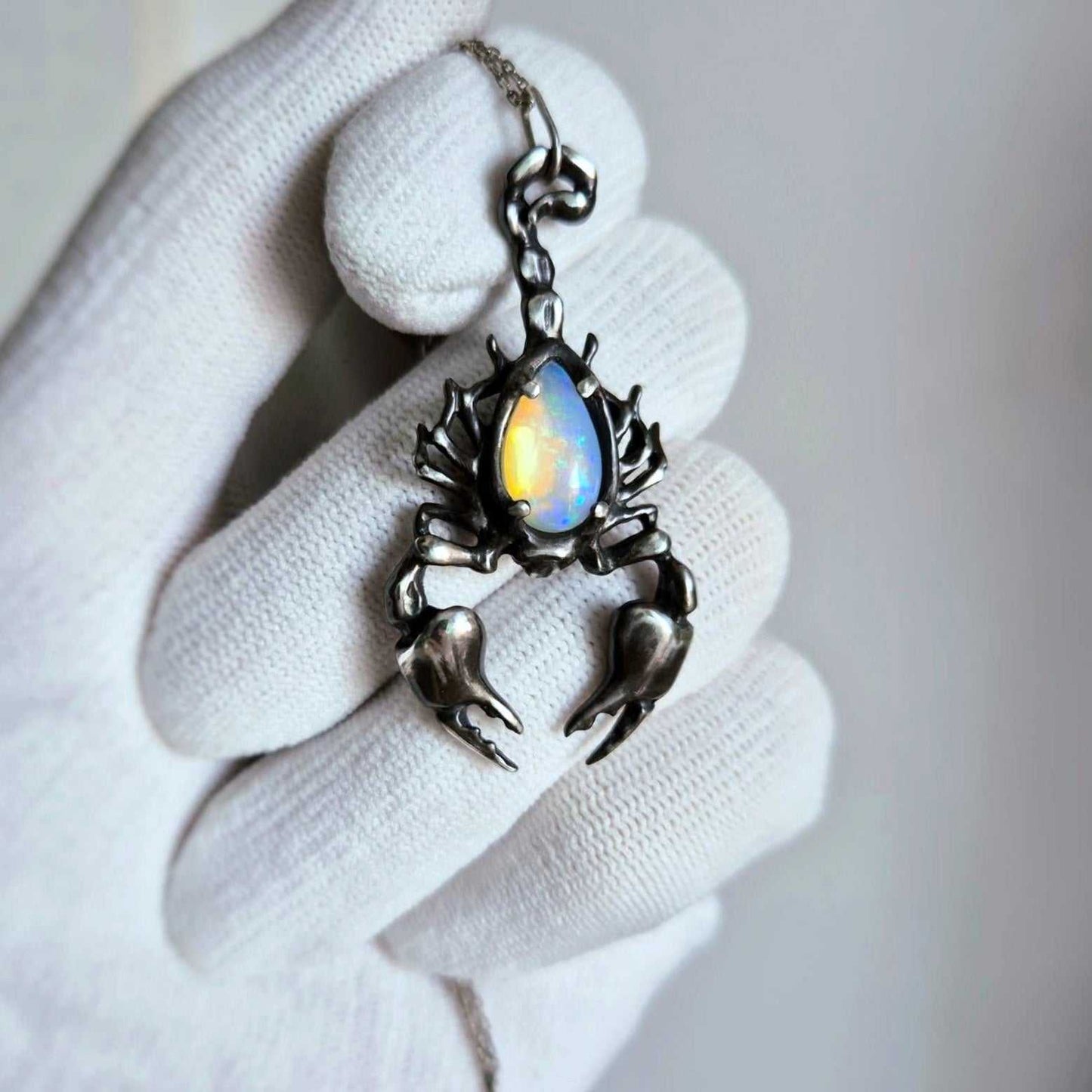 Close-up of a silver Scorpion necklace with a luminous opal gemstone, held in a white-gloved hand