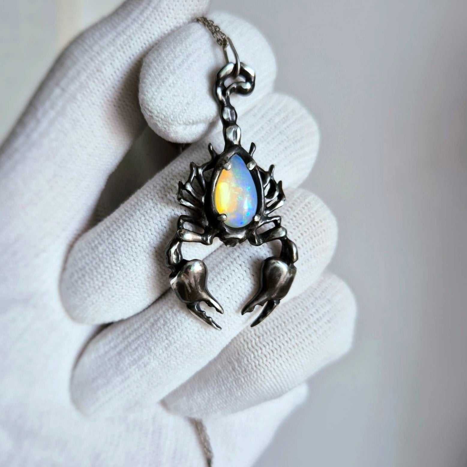 Close-up of a silver Scorpion necklace with a luminous opal gemstone, held in a white-gloved hand