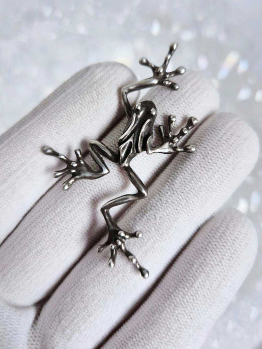 Full silver frog pendant held in a white-gloved hand, showing its long legs and splayed toes