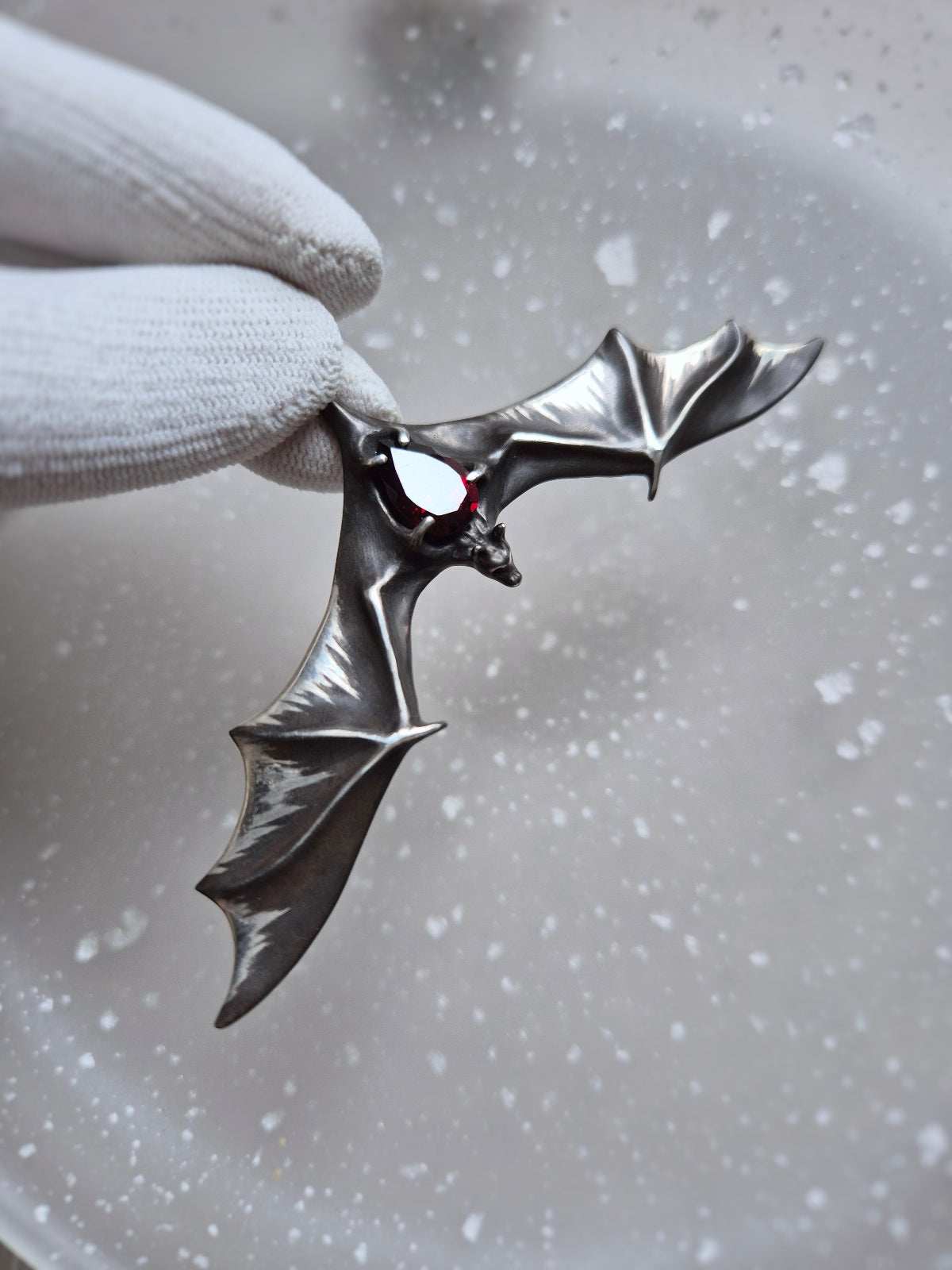 Silver Bat necklace with wide sculpted wings and a natural garnet, held in a gloved hand.