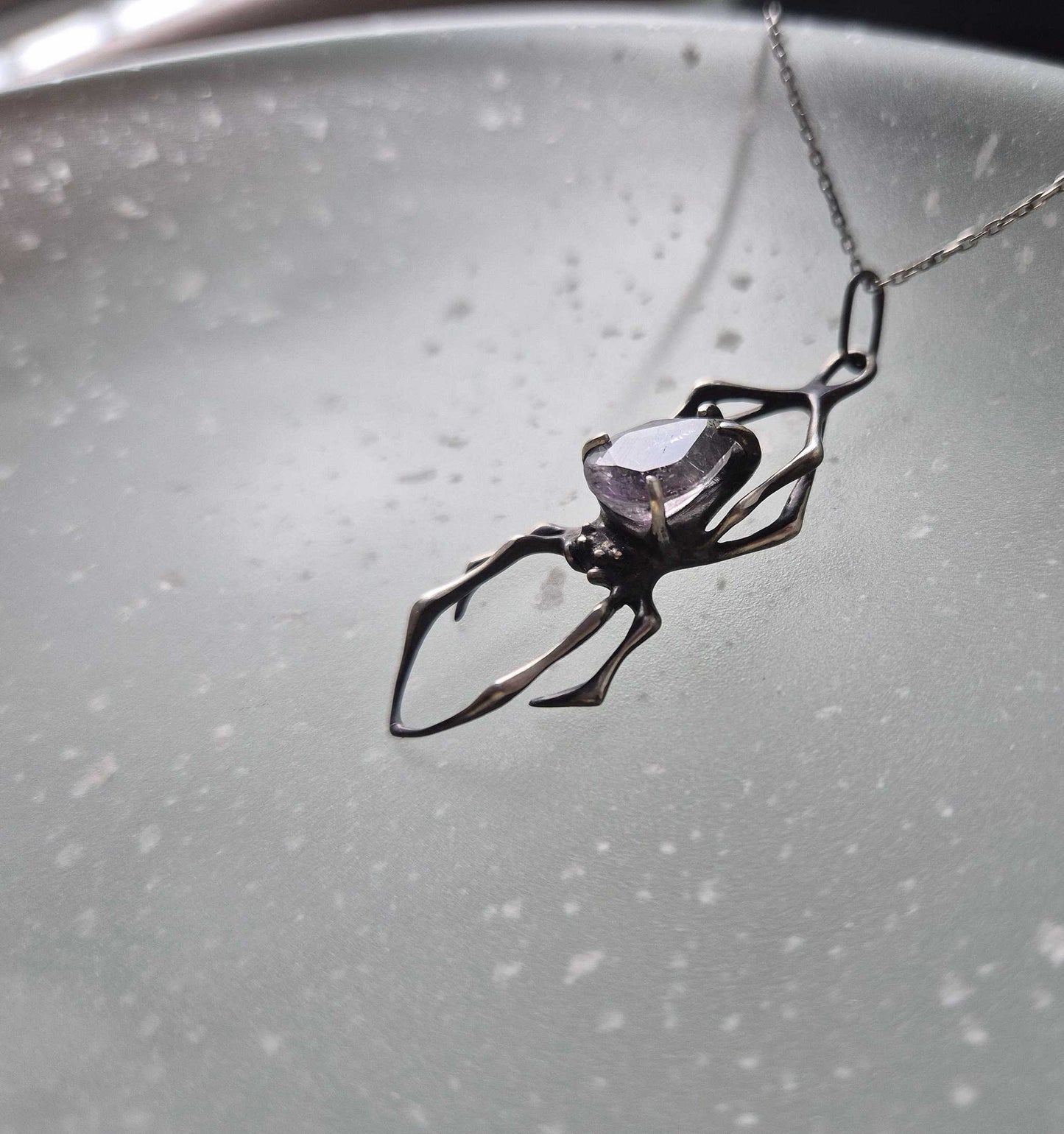 Side view of a silver spider pendant with super seven quartz gem, on a glass surface with soft speckled light patterns
