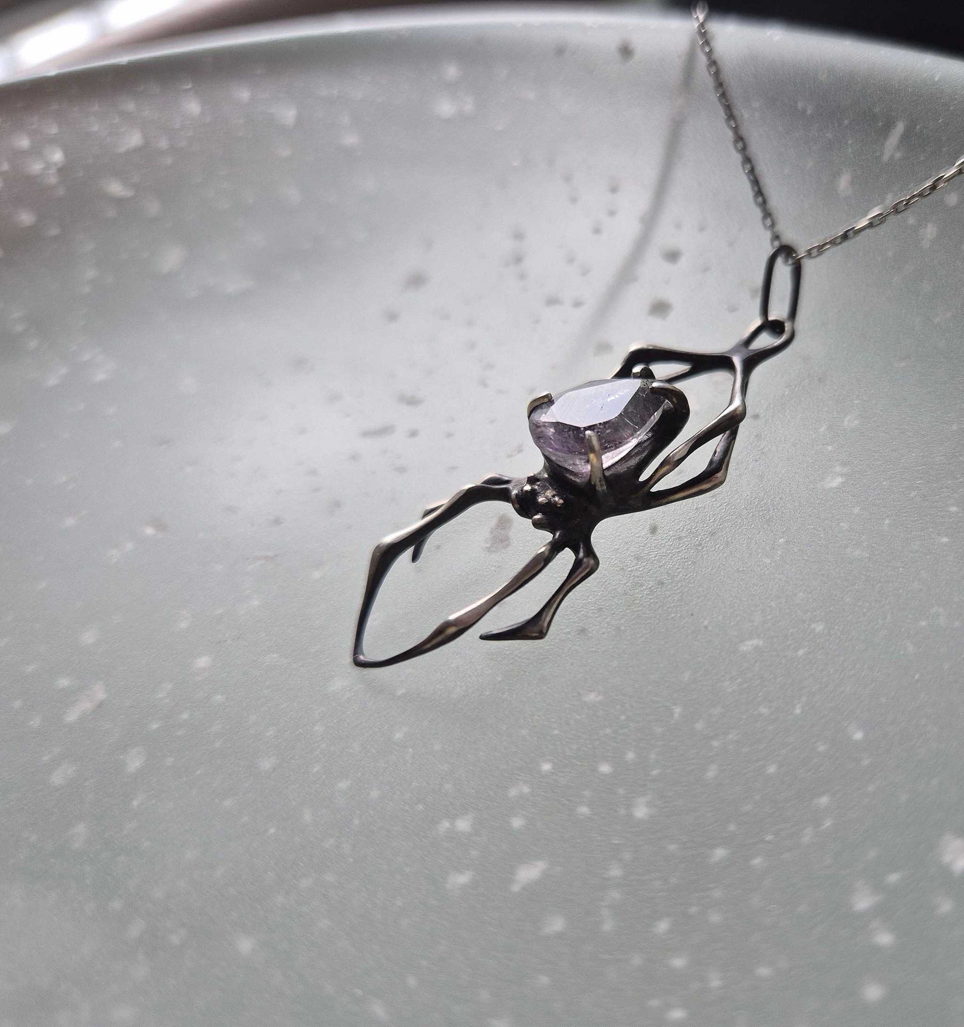 Side view of a silver spider pendant with super seven quartz gem, on a glass surface with soft speckled light patterns