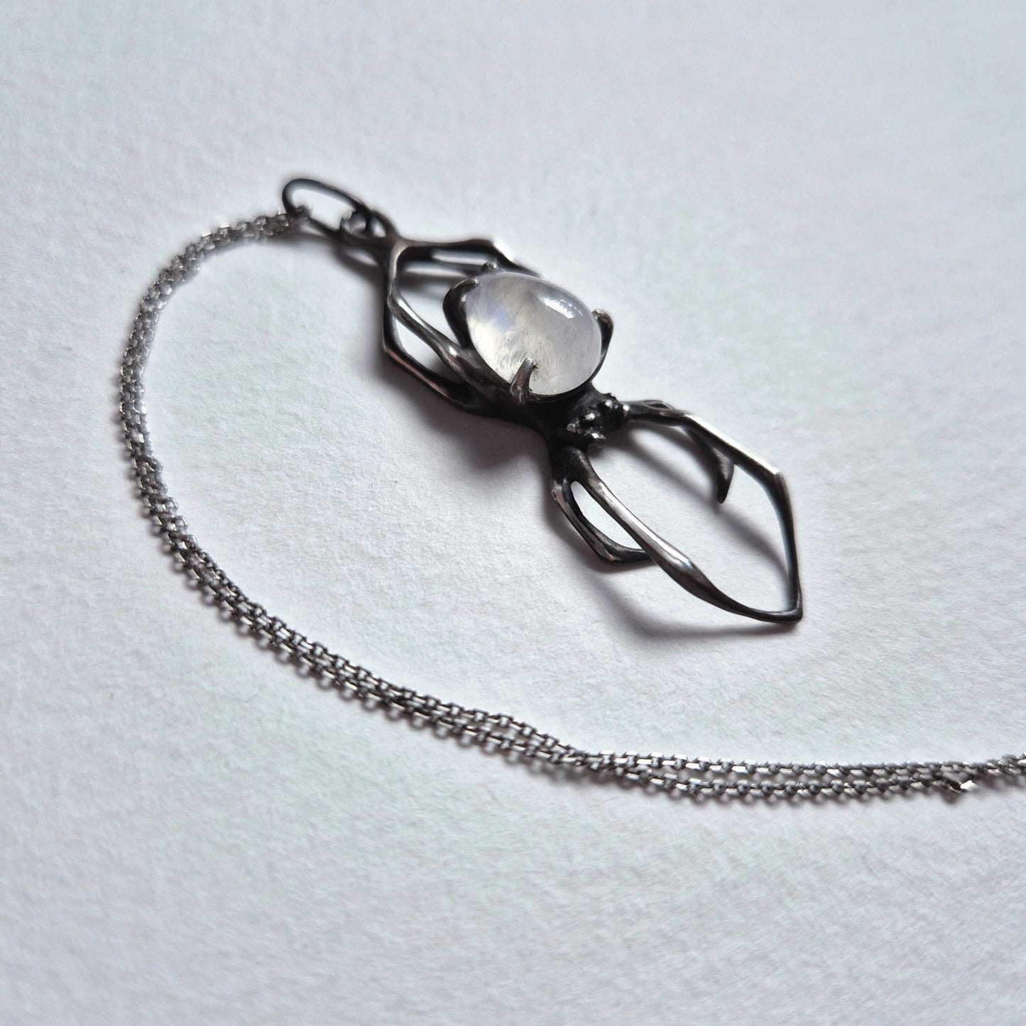 Side view of silver spider necklace with moonstone lying on white surface, highlighting details and gentle curve of the legs