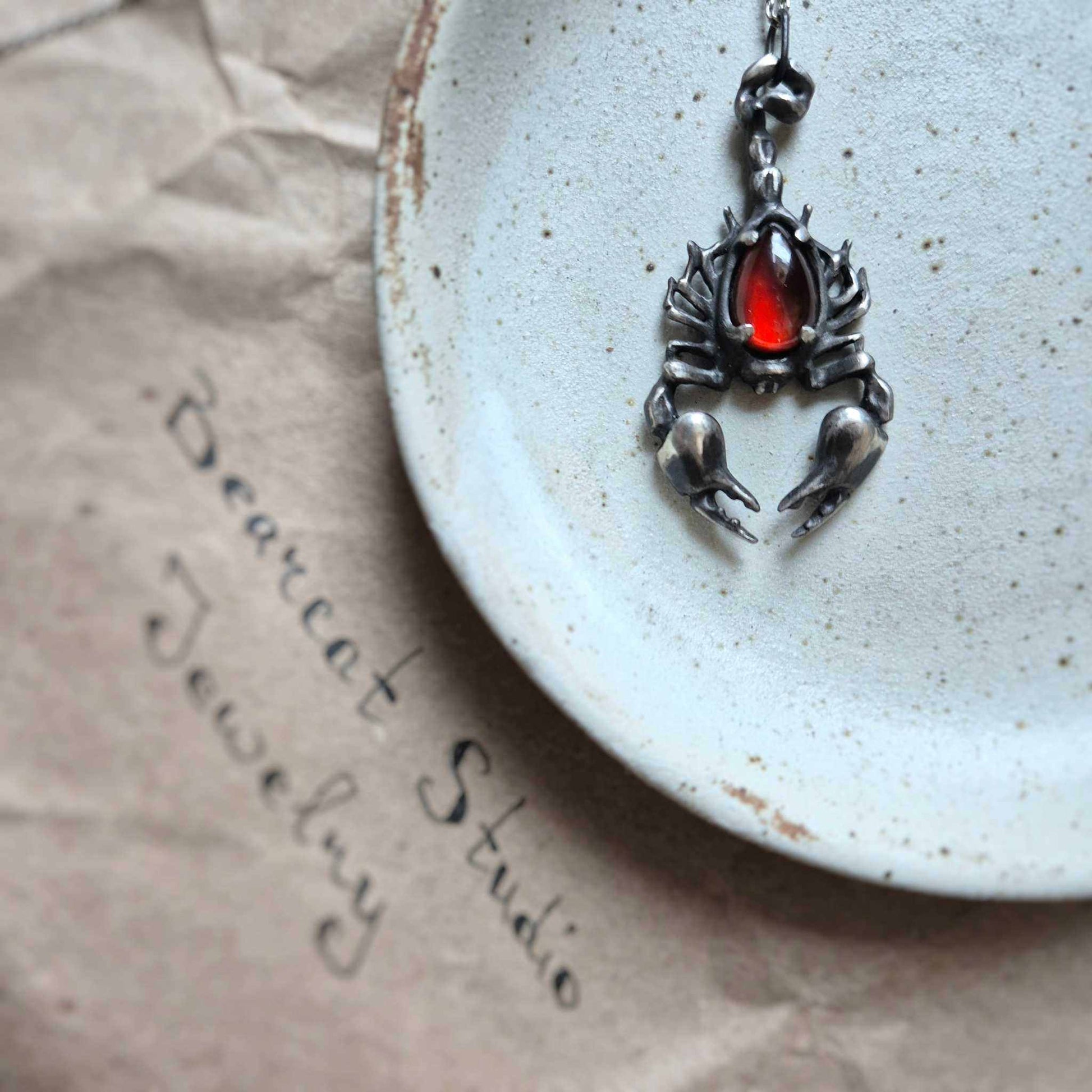 Close-up of a silver Scorpion necklace with a deep red gemstone, placed on a speckled ceramic plate with the handwritten Bearcat Studio Jewelry branding.