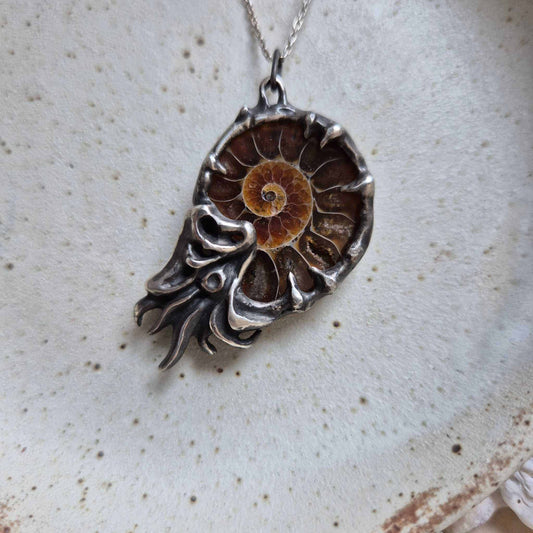 Close-up of a silver Nautilus necklace with a natural ammonite fossil, lying on a softly speckled ceramic plate