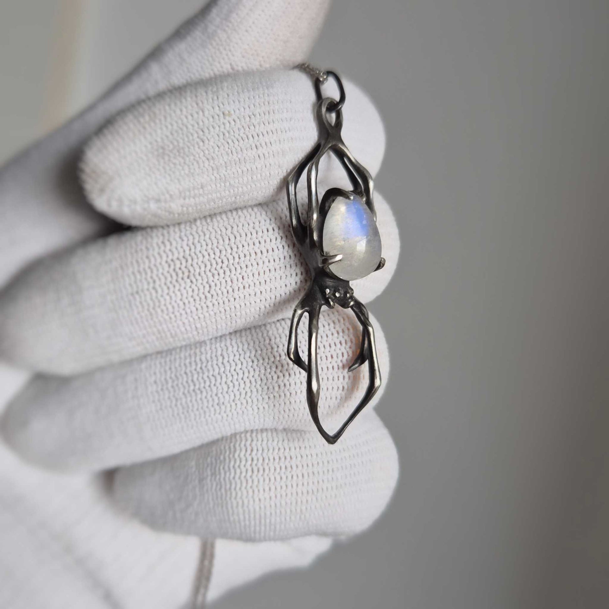Gloved hand holding silver spider pendant with rainbow moonstone cabochon