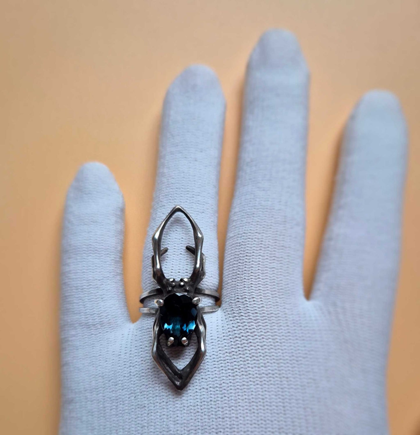 Silver spider ring with London blue topaz shown on white gloved hand, orange background