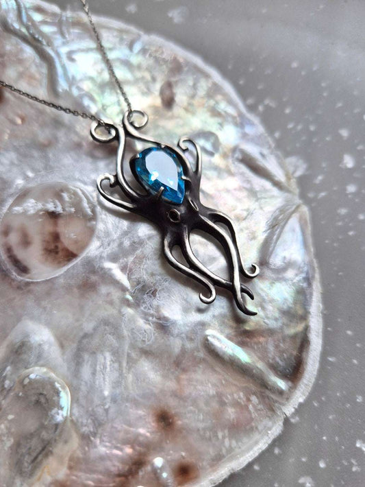 Silver Octopus necklace with teardrop-cut blue gemstone, displayed on iridescent shell surface